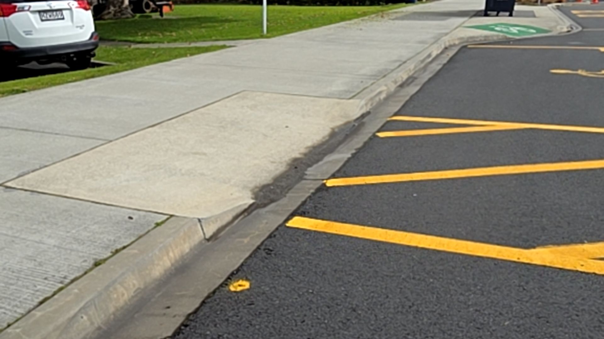 Sloped concrete kerb ramp connecting a level grey footpath to an asphalt parking area with yellow hatched markings, providing a transition between the raised path and road level.