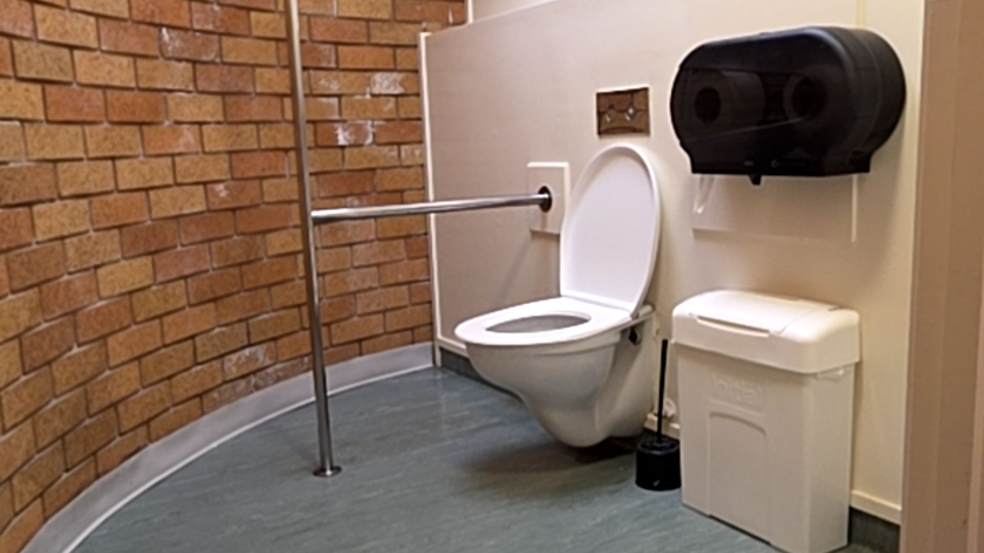 White wall-hung toilet with T-shaped stainless steel grab rail. Level grey flooring with clear space. Curved brick wall and beige partition with a black toilet paper dispenser and white bin.