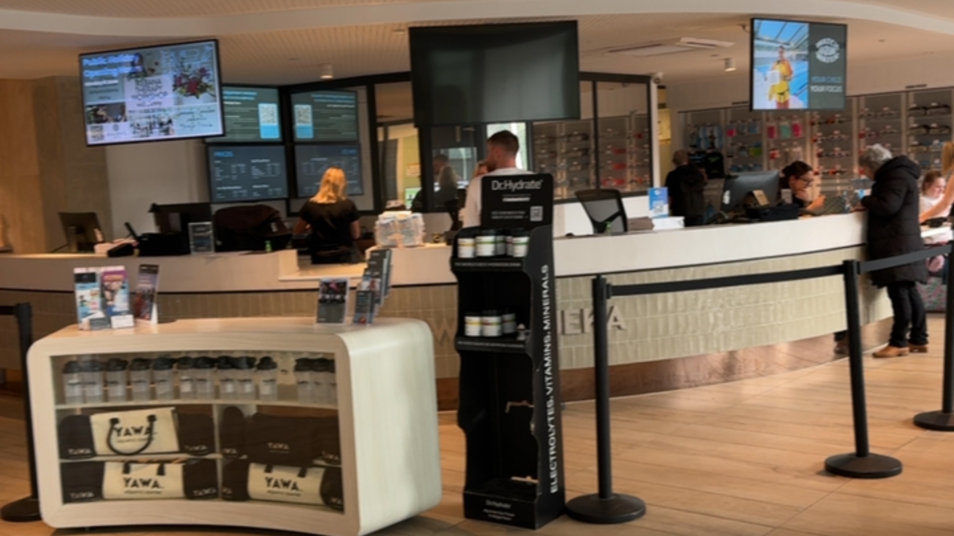 Bright centre reception with smooth, level tiled floor. Long white service counter with black belt stanchions. Wide, unobstructed pathways around low-profile merchandise display units.