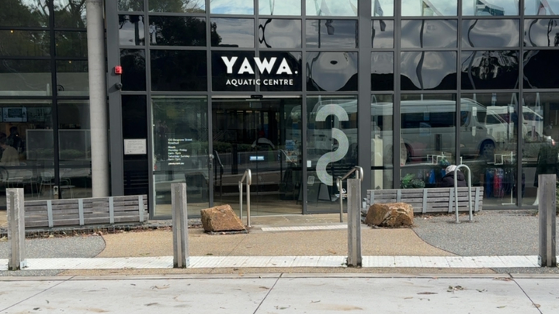 Level entrance to Yawa Aquatic Centre via automatic glass doors. Tan textured pathway with white tactile strips and silver handrails. Flanked by timber bollards and low timber bench seating.
