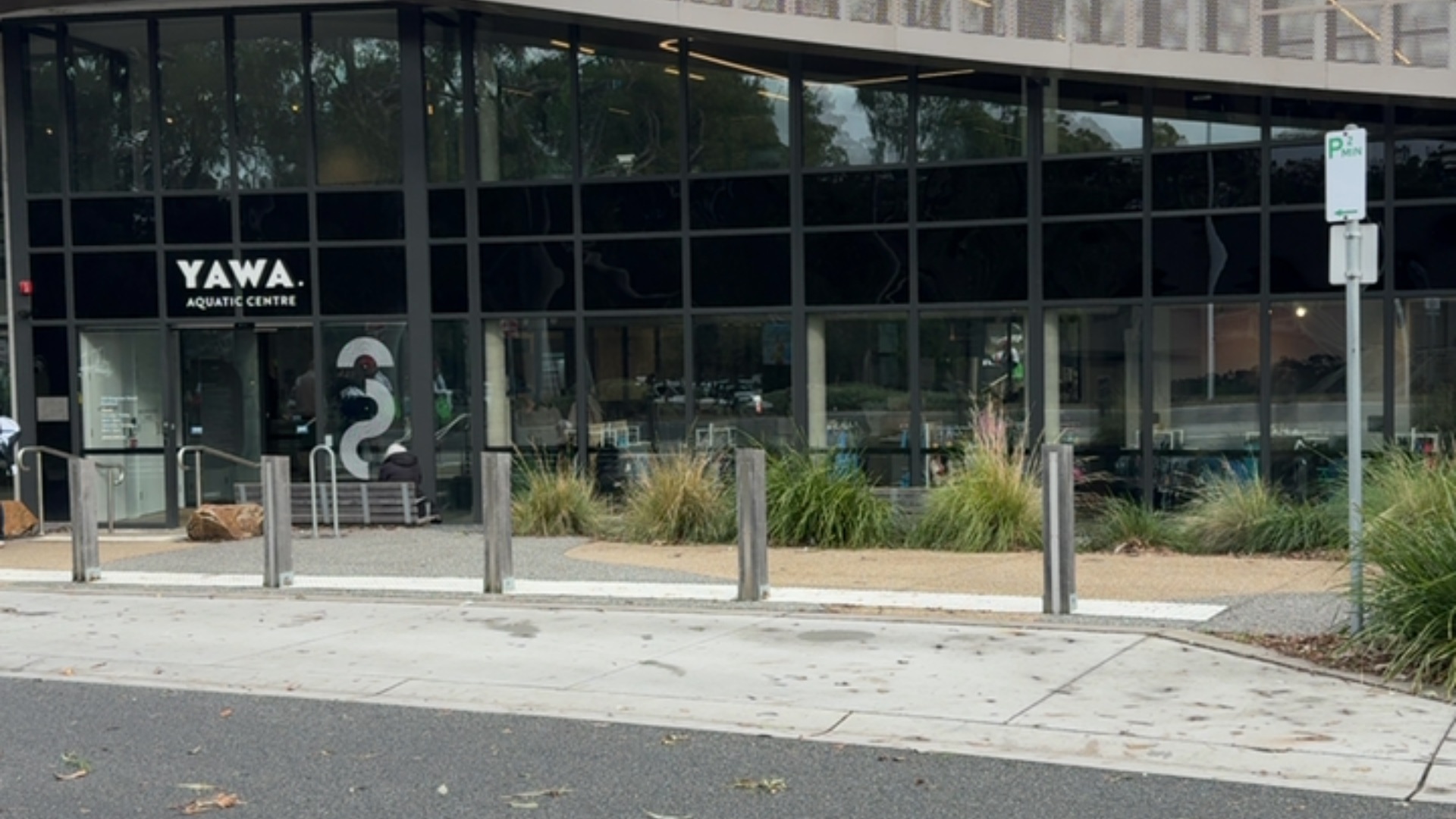 Wide level concrete path leads to YAWA Aquatic Centre ground-level glass sliding doors. Timber bollards line the path near a timber bench and metal handrail. The entry area is flat.