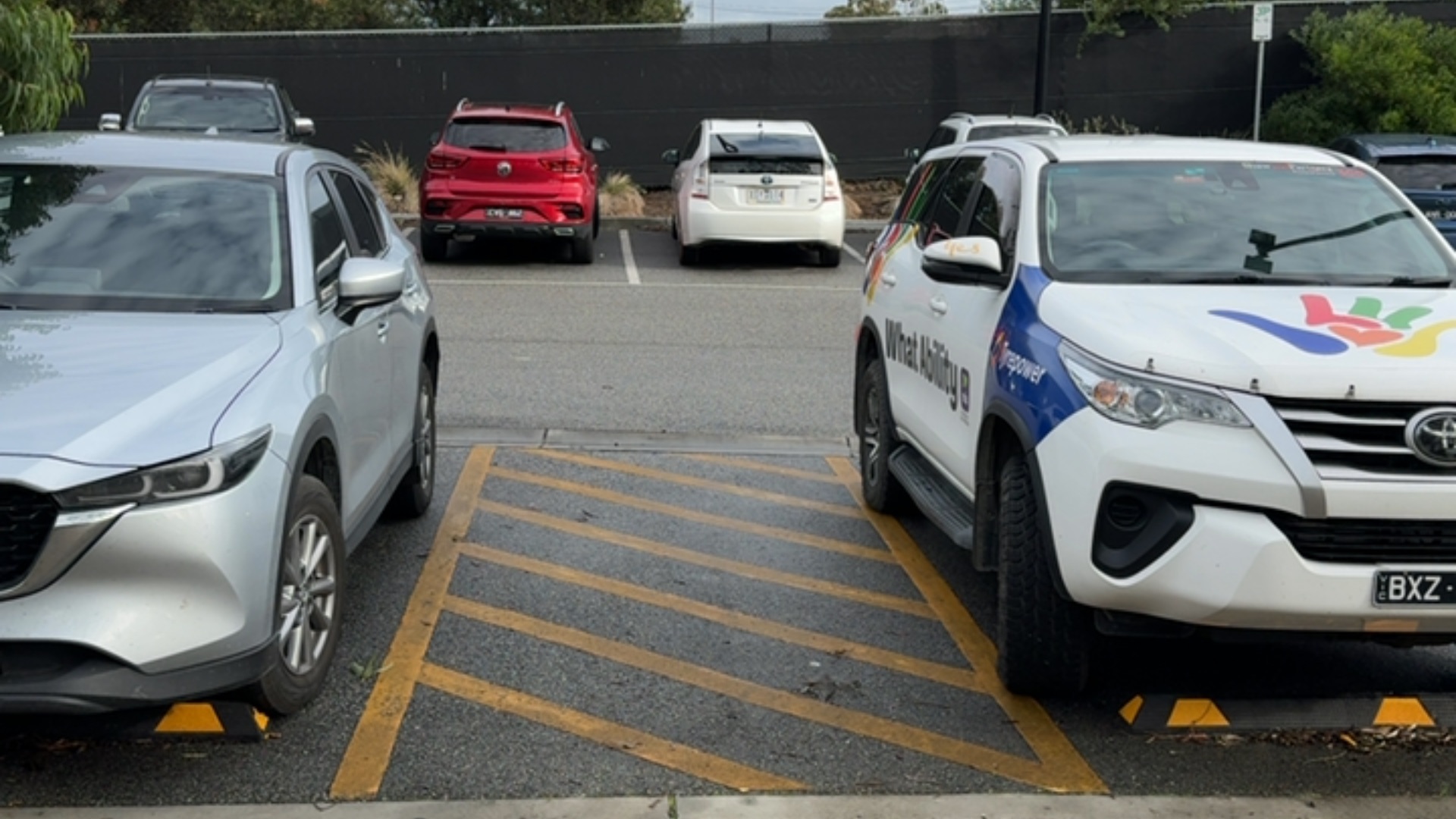Yellow diagonal-lined shared zone on level asphalt between a silver SUV and a white SUV. This access aisle provides extra width for mobility aids between the two vehicles.