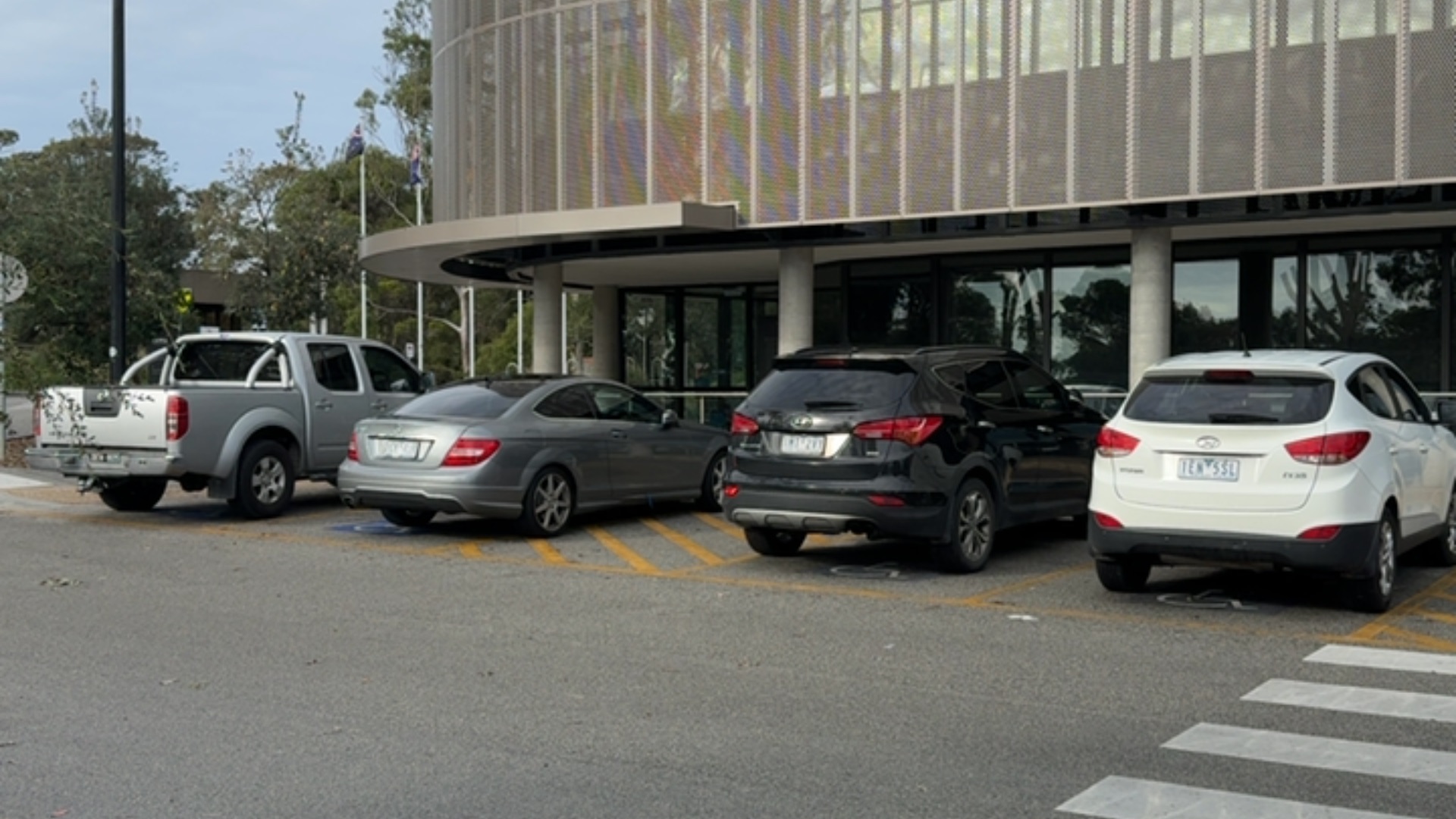Level bitumen car park with yellow hatched accessible bays and wheelchair symbols. A white pedestrian crossing on the right leads to a modern building with glass door entrances.