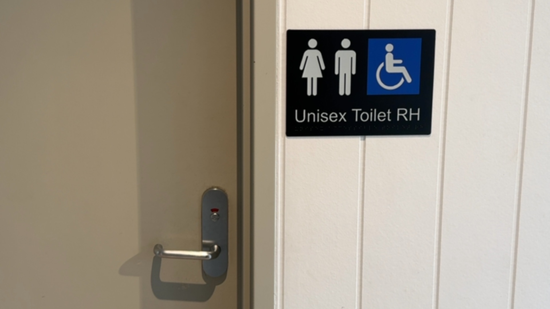 Light-grey door with silver lever handle and red indicator. Beside it, a black sign with white male, female and blue wheelchair icons, text 'Unisex Toilet RH' and braille.