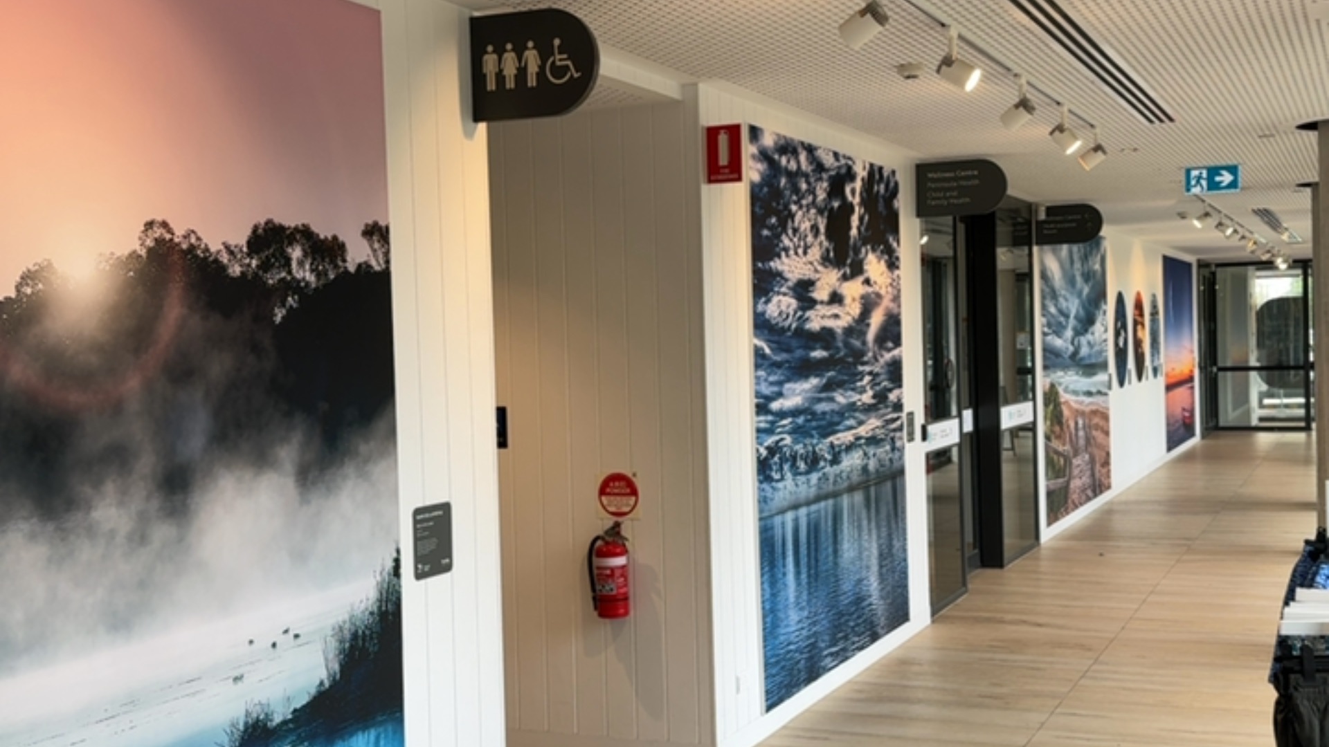 Wide, level hallway with timber-look flooring. Left entrance with accessible toilet signage. Well-lit corridor features Wellness Centre signage and a green emergency exit sign at the end.