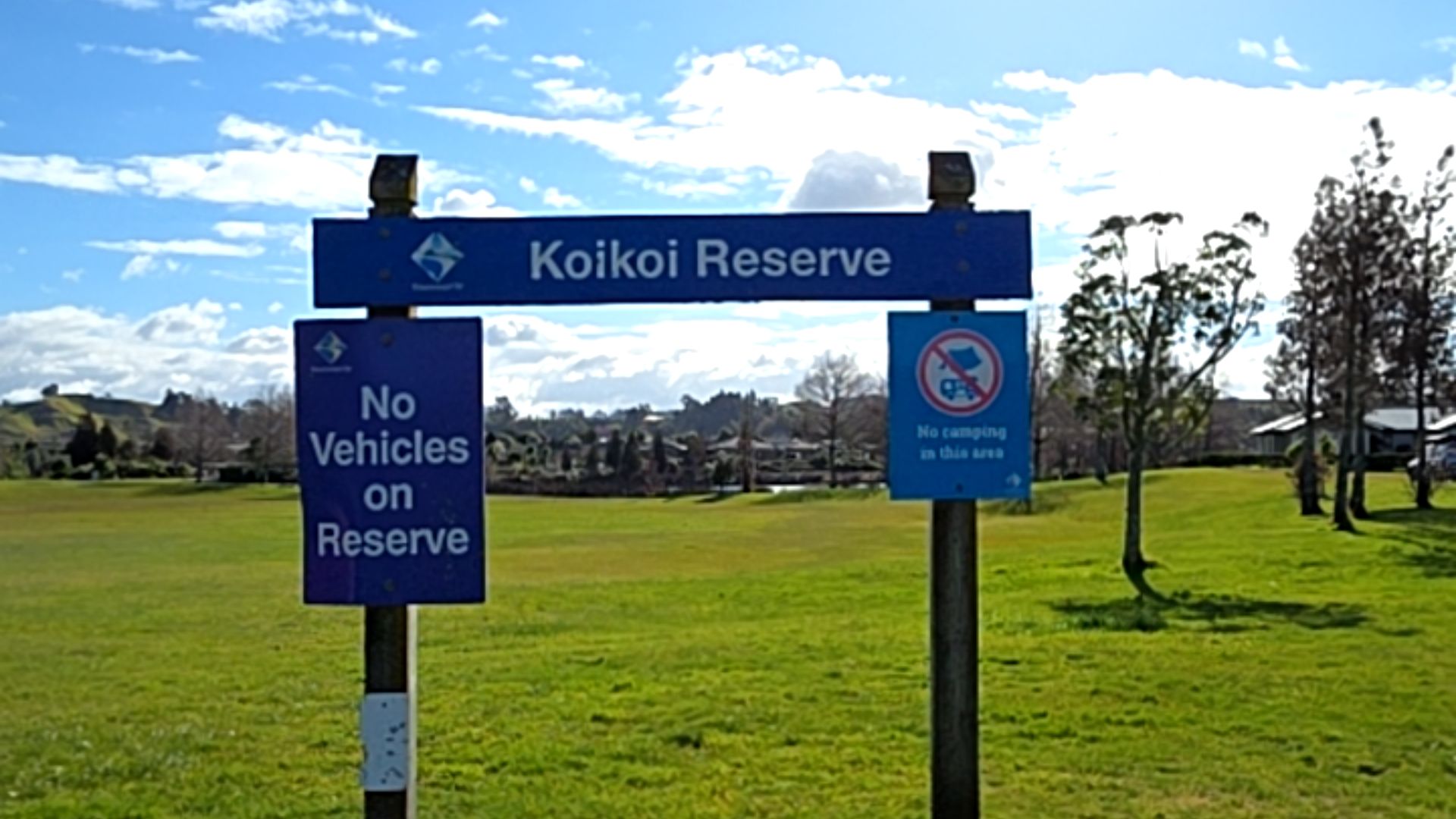 Blue 'Koikoi Reserve' sign on timber posts. Attached signs read 'No Vehicles on Reserve' and 'No camping in this area' with symbols. Level grass parkland with trees in the background.