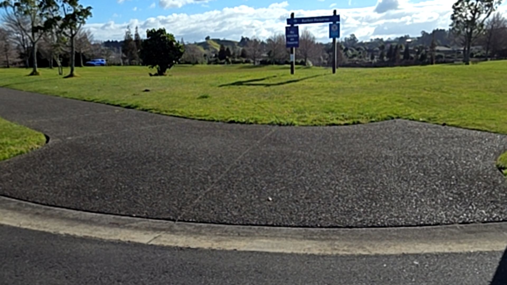 Wide dark grey asphalt pathway starts at a level concrete kerb, leading into an open grassy park. A tall blue signpost stands on the lawn in the background.