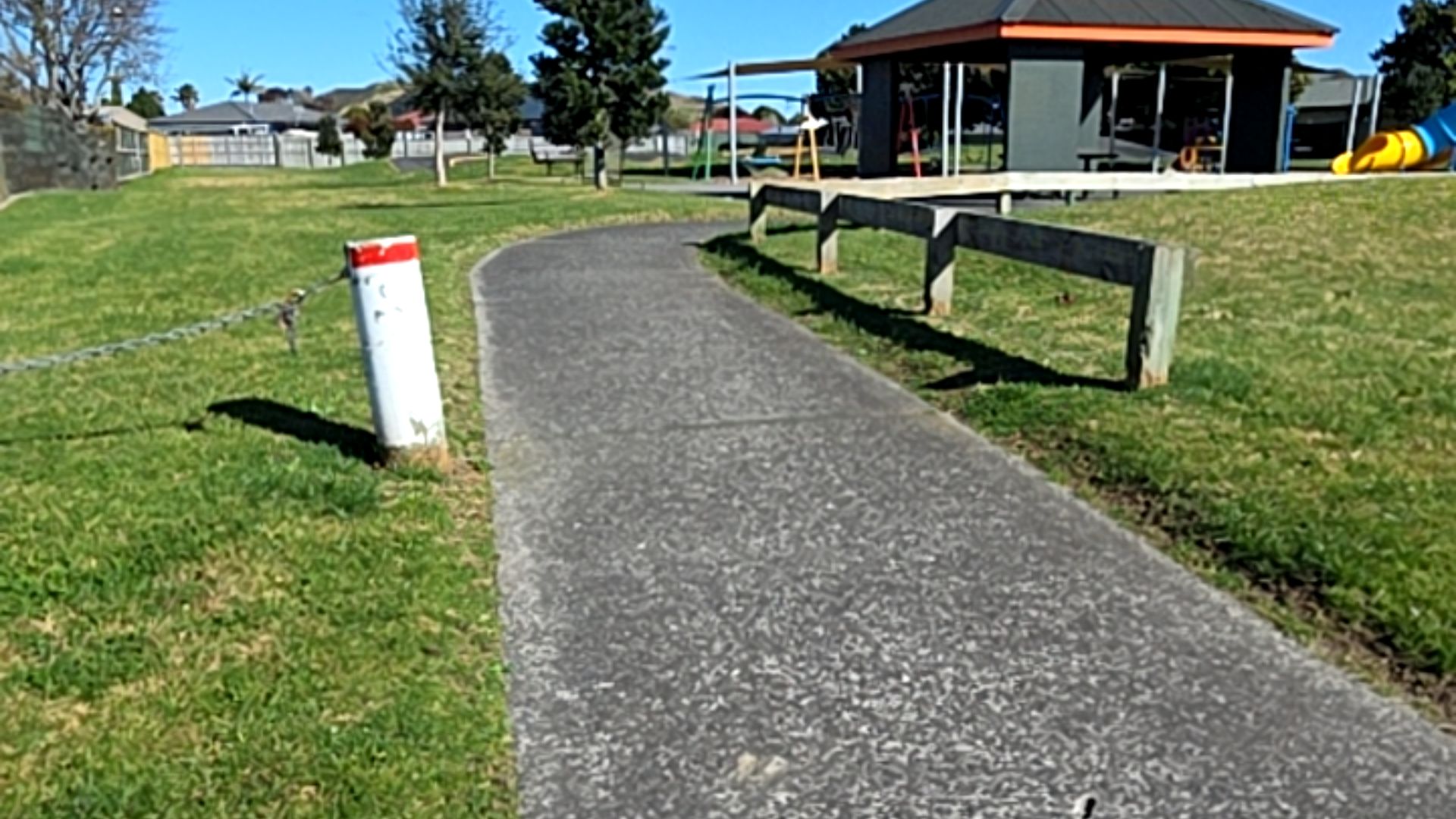 Level asphalt path curves through grass. Left: white bollard with red band and chain. Right: low timber rail fence. Background: grey shelter and colourful playground.