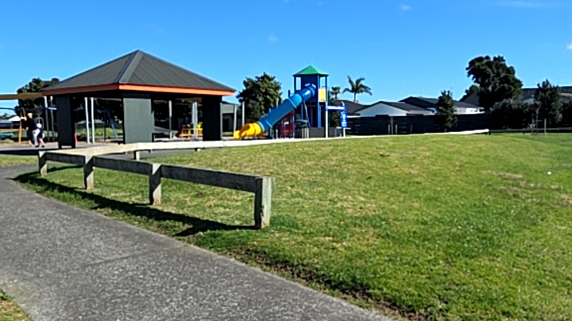 Wide, level concrete pathway beside a low timber post-and-rail fence. Flat grassed area leads to a grey open-sided shelter and a playground with a blue and yellow slide.