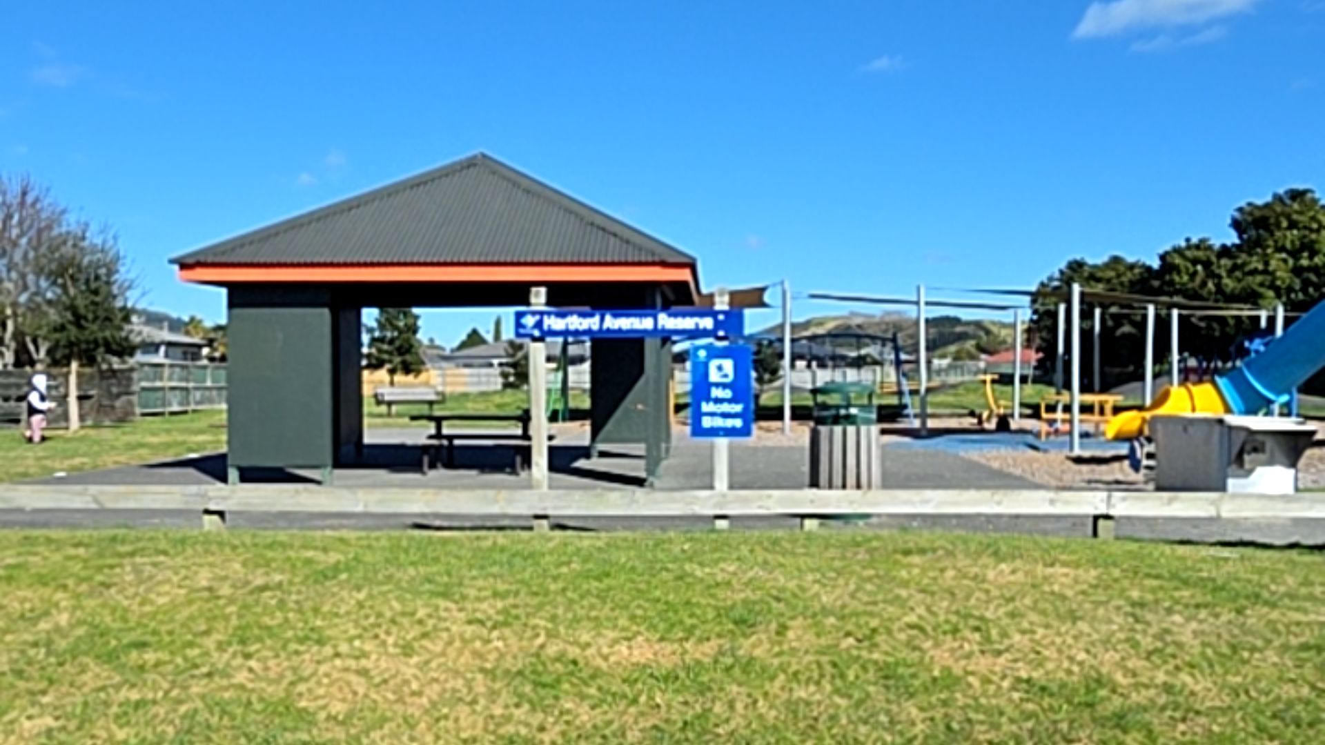 Blue signs for Hartford Avenue Reserve and No Motor Bikes. Picnic shelter with table on a level grey surface. Low timber fence separates grass foreground from playground area in background.