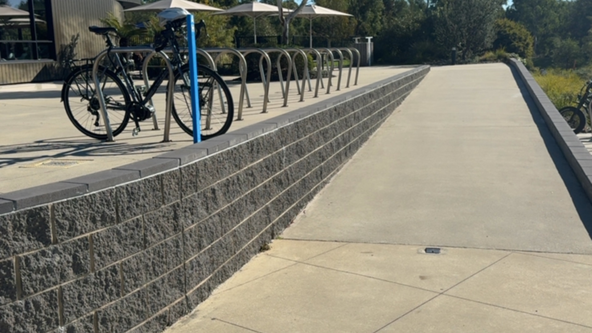 Wide concrete ramp with gentle gradient beside a low charcoal grey textured stone wall with smooth capping. Silver metal hoop bike racks are on the level concrete area at the top.