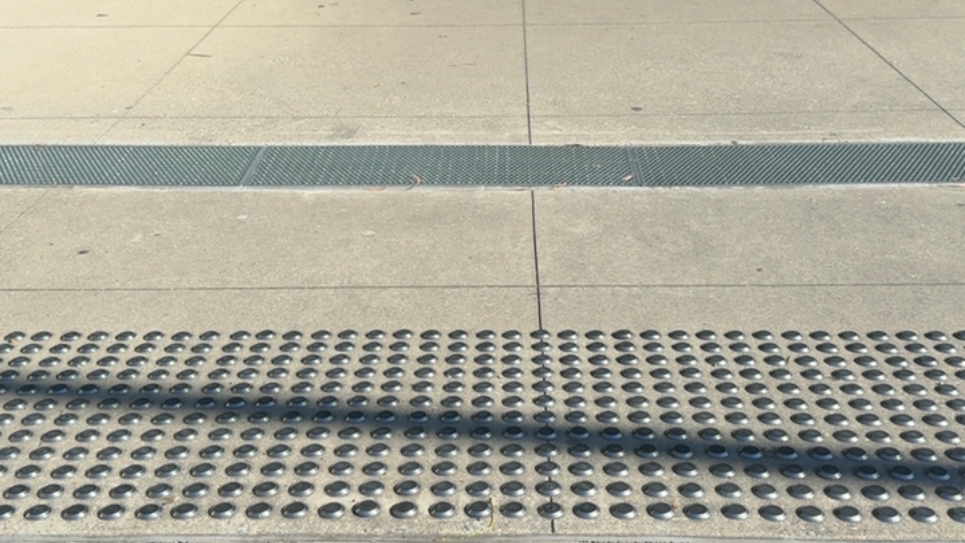 Level concrete pavement with a strip of dark grey metal grating and a section of raised circular tactile paving studs in a grid pattern.