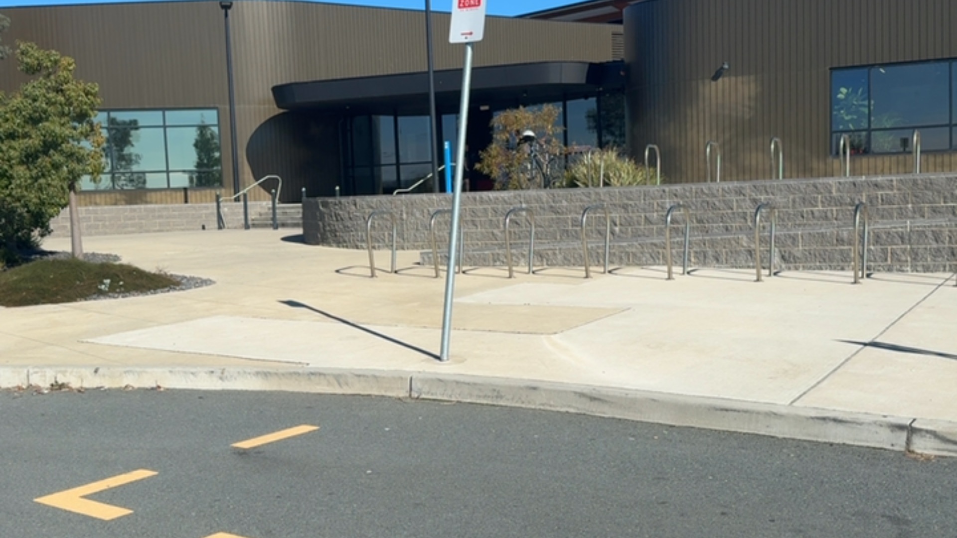 Level concrete pathway leads to a glass entrance. A ramp with metal handrails rises behind U-shaped bike racks. A sign on a tall pole stands near the asphalt road and concrete kerb.