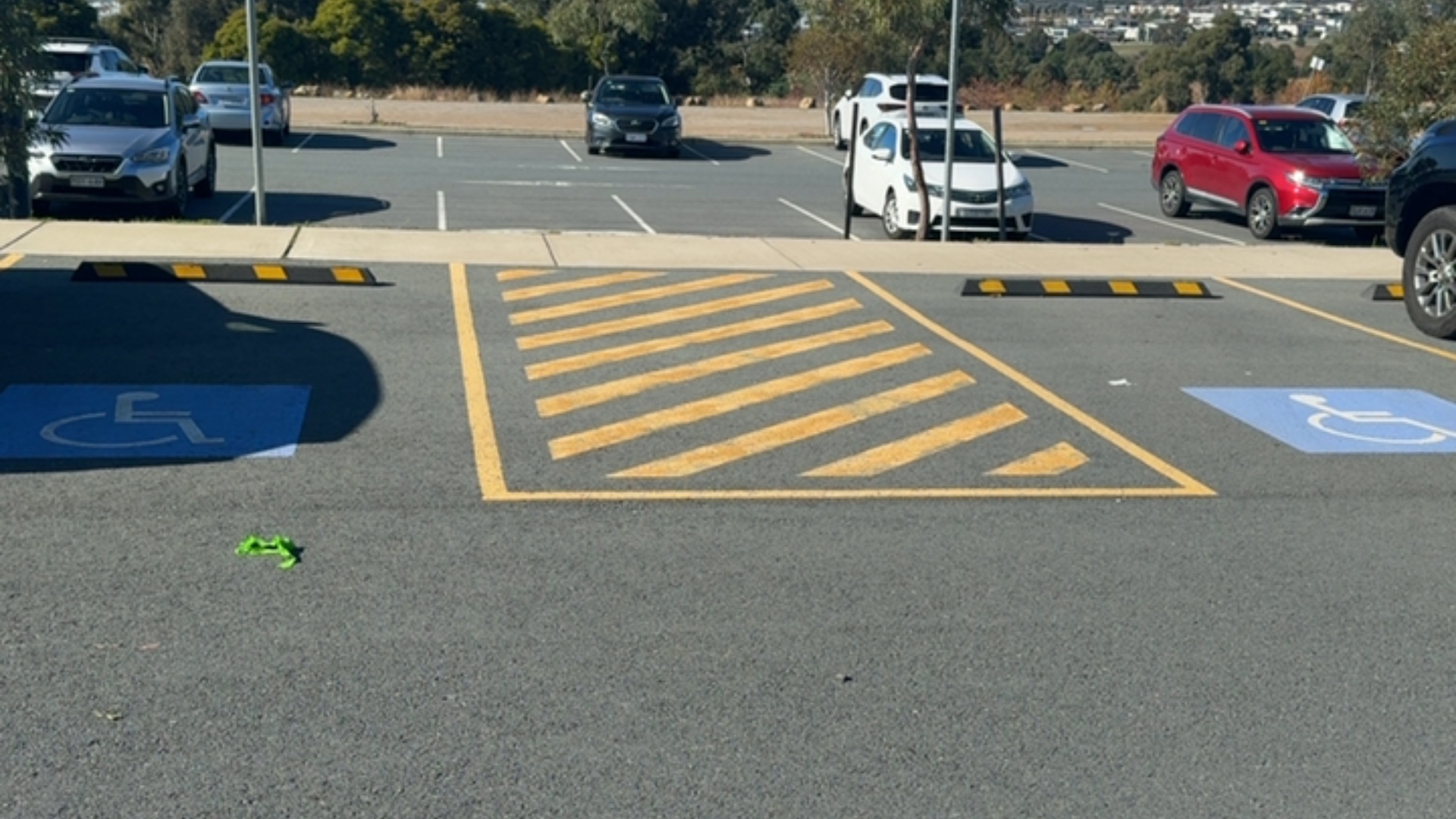 Two level accessible parking bays with blue wheelchair symbols on asphalt. A yellow cross-hatched shared access zone sits between them, with black and yellow wheel stops along the concrete kerb.