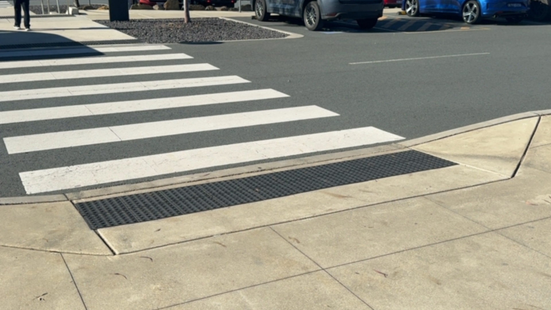 Level concrete footpath with black tactile ground surface indicators leading to a sloped kerb ramp and a white-striped zebra crossing on a dark grey bitumen road surface.