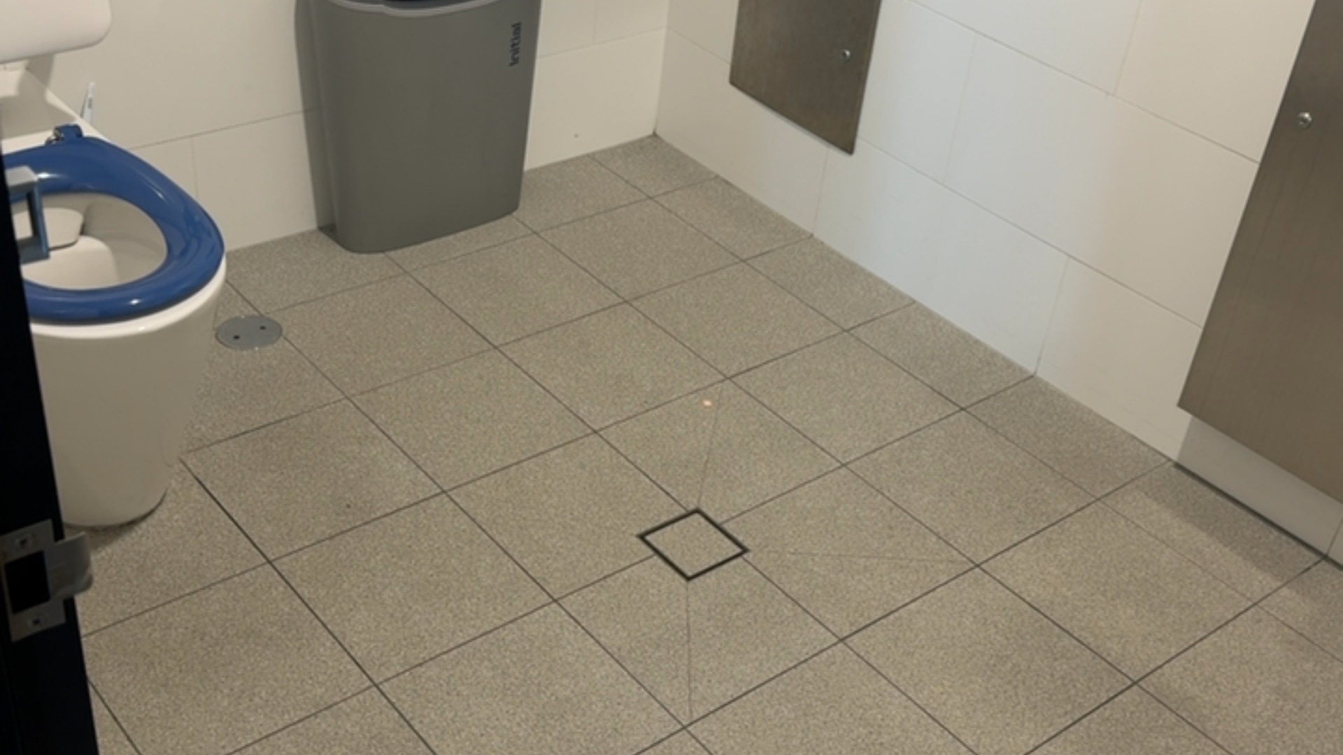 Level grey tiled floor in a toilet cubicle with clear floor space. White toilet with a high-contrast blue seat. Central square drain and a tall grey bin in the corner.