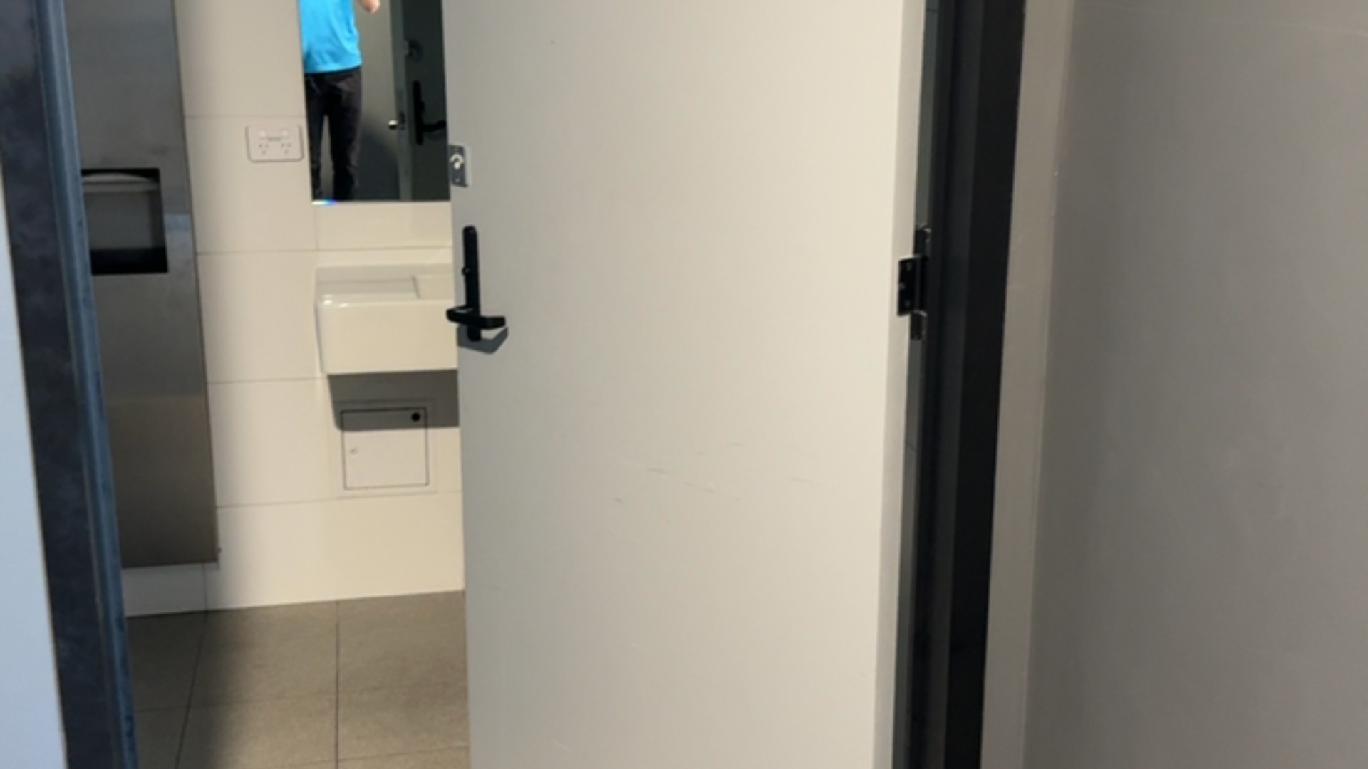 Wide white door with a black lever handle opens onto a level entrance with grey floor tiles. Inside features a wall-mounted basin and mirror.