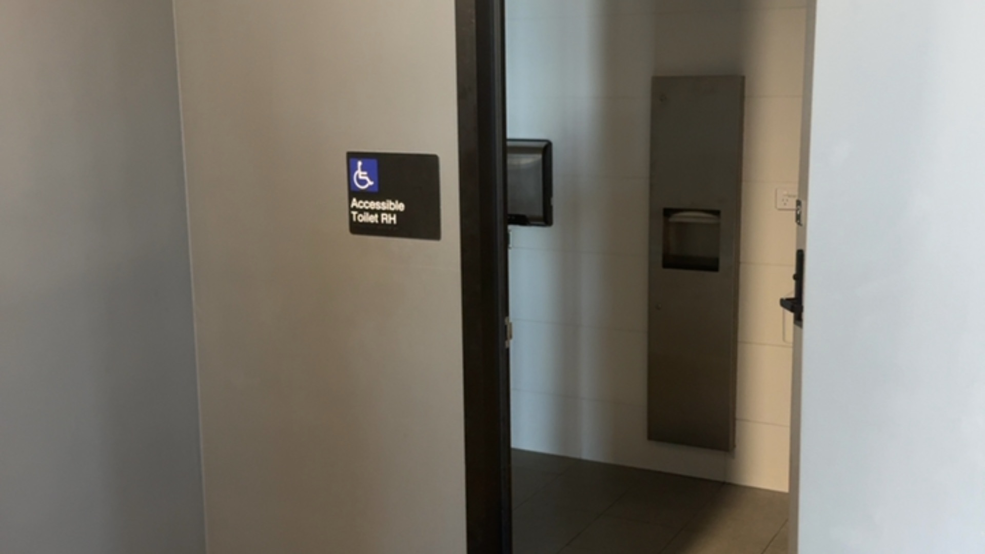 Accessible toilet entrance with a black wall sign featuring a blue wheelchair symbol and text 'Accessible Toilet RH'. Level entrance leads to tiled flooring and a recessed dispenser.