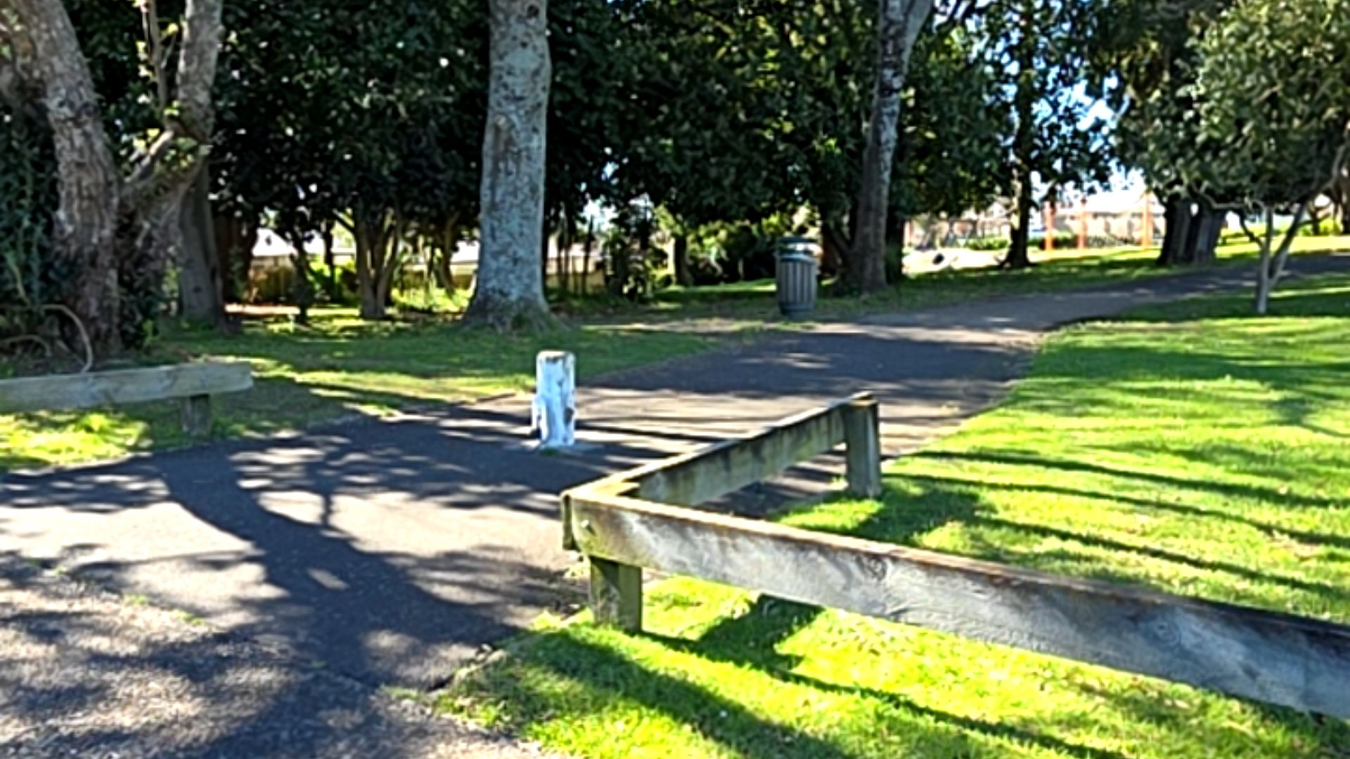Level asphalt path with a central white bollard, bordered by low wooden post and rail fencing. Grassy areas and large trees surround the area, with a grey rubbish bin in the background.