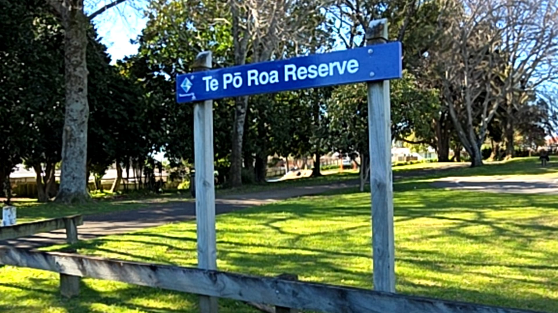Blue sign on grey wooden posts reads 'Te Pō Roa Reserve' in white. A level, dark-coloured paved pathway leads through a grassy park with large shady trees and a low wooden fence.