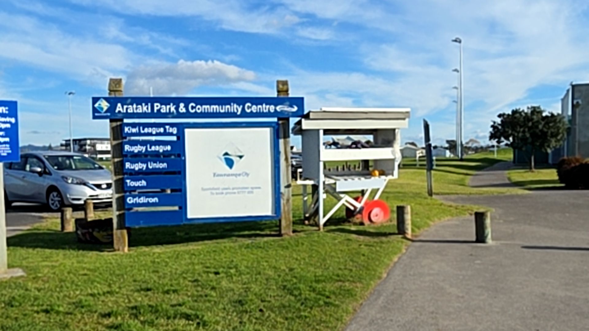 Level bitumen path with wooden bollards. Blue Arataki Park & Community Centre sign on grass. Small white cart with red wheels. Level asphalt parking area and silver car in background.