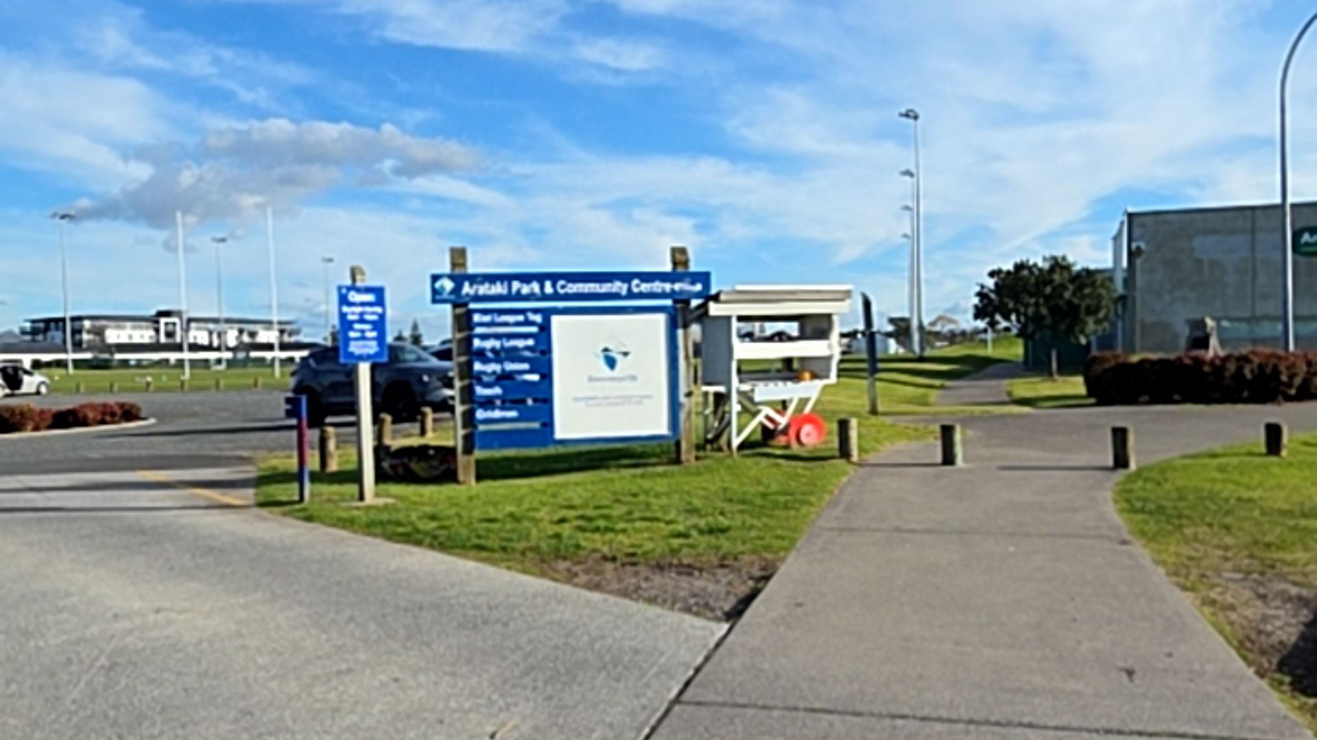 Arataki Park & Community Centre sign beside a wide, level concrete pathway. Flat asphalt car park and grass area with wooden bollards. Level ground throughout.