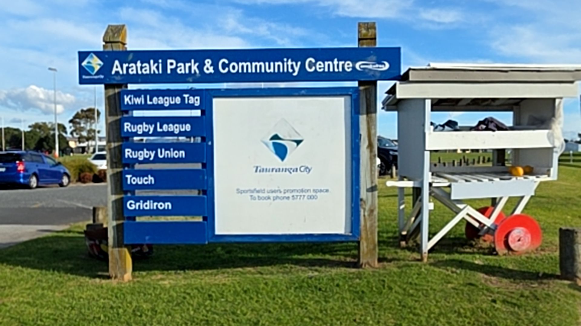 Blue sign for Arataki Park & Community Centre listing sports. It stands on wooden posts in grass beside an asphalt road. A white wooden cart with red wheels sits to the right.