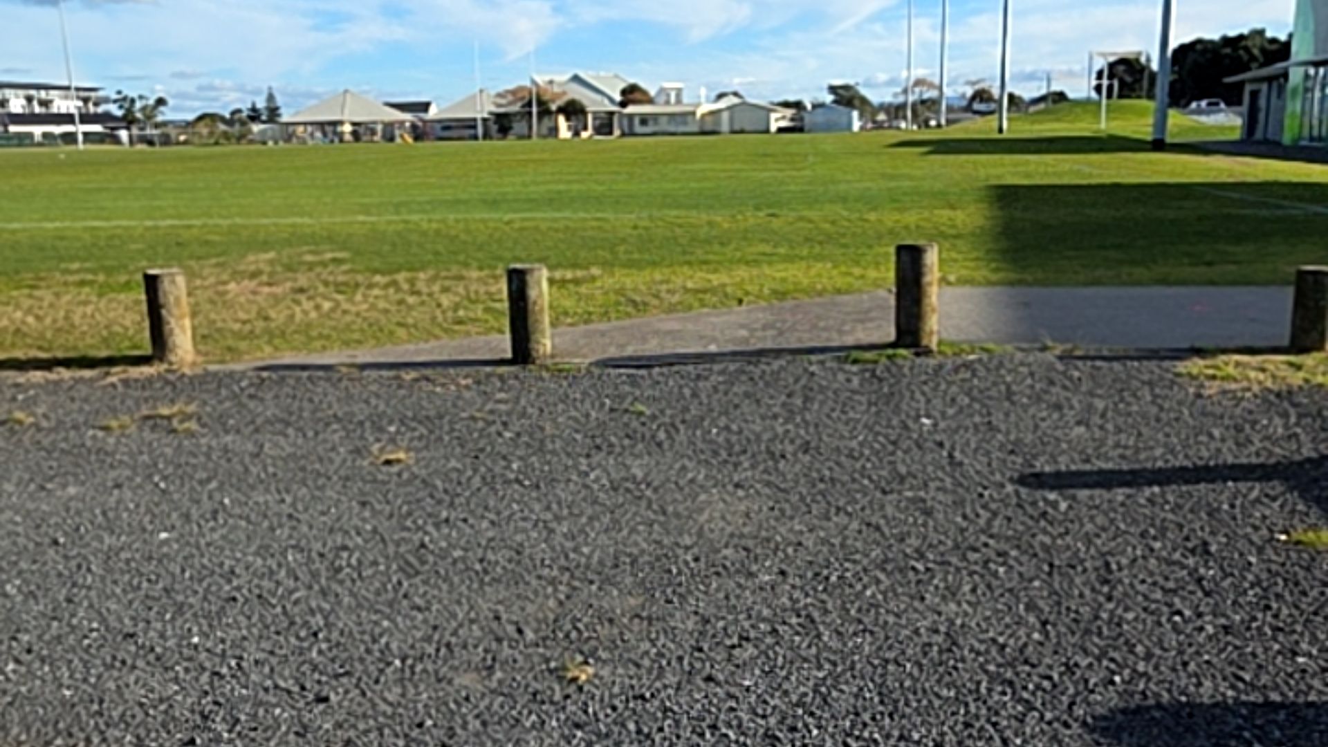 Level gravel area meets a flat concrete pathway separated by four wooden bollards. Beyond is a large, level green sports field with tall light poles and distant houses.