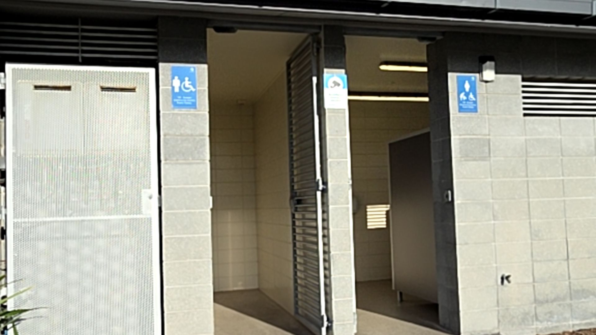 Level concrete block toilet entrances with blue signs. Left: male and wheelchair icons. Right: open metal gate, female, child and wheelchair icons. Tiled floors and bright internal lighting.