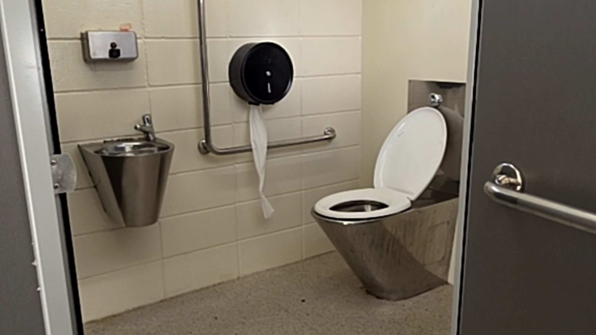 Stainless steel toilet with white seat and L-shaped grab rail. Wall-mounted basin and soap dispenser. Cream tiled walls, grey non-slip floor, and dark grey door with horizontal metal rail.