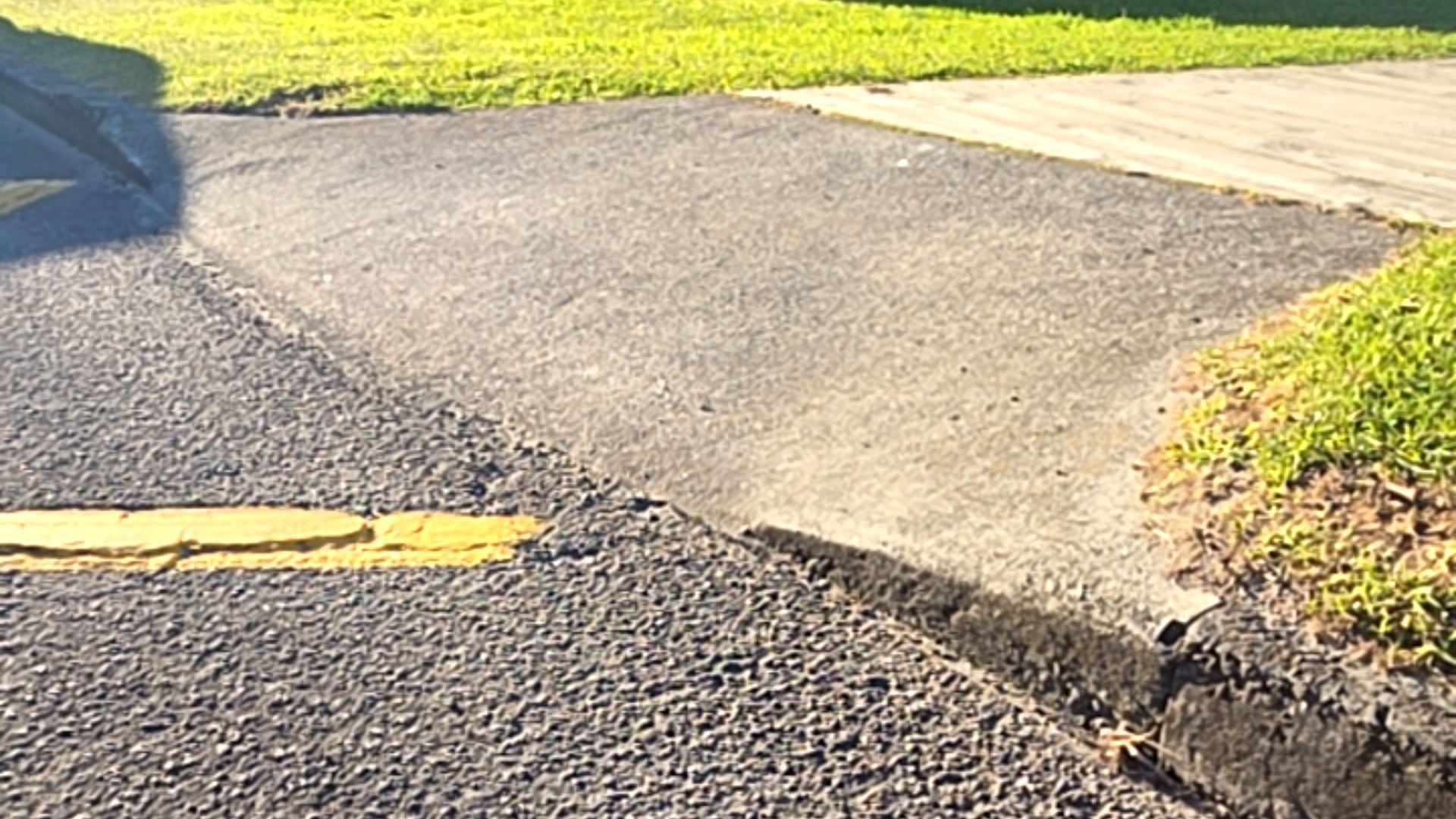 Concrete driveway ramp meets a dark asphalt road with a yellow line. An uneven join creates a vertical lip between the surfaces. Grass borders the concrete pathway.