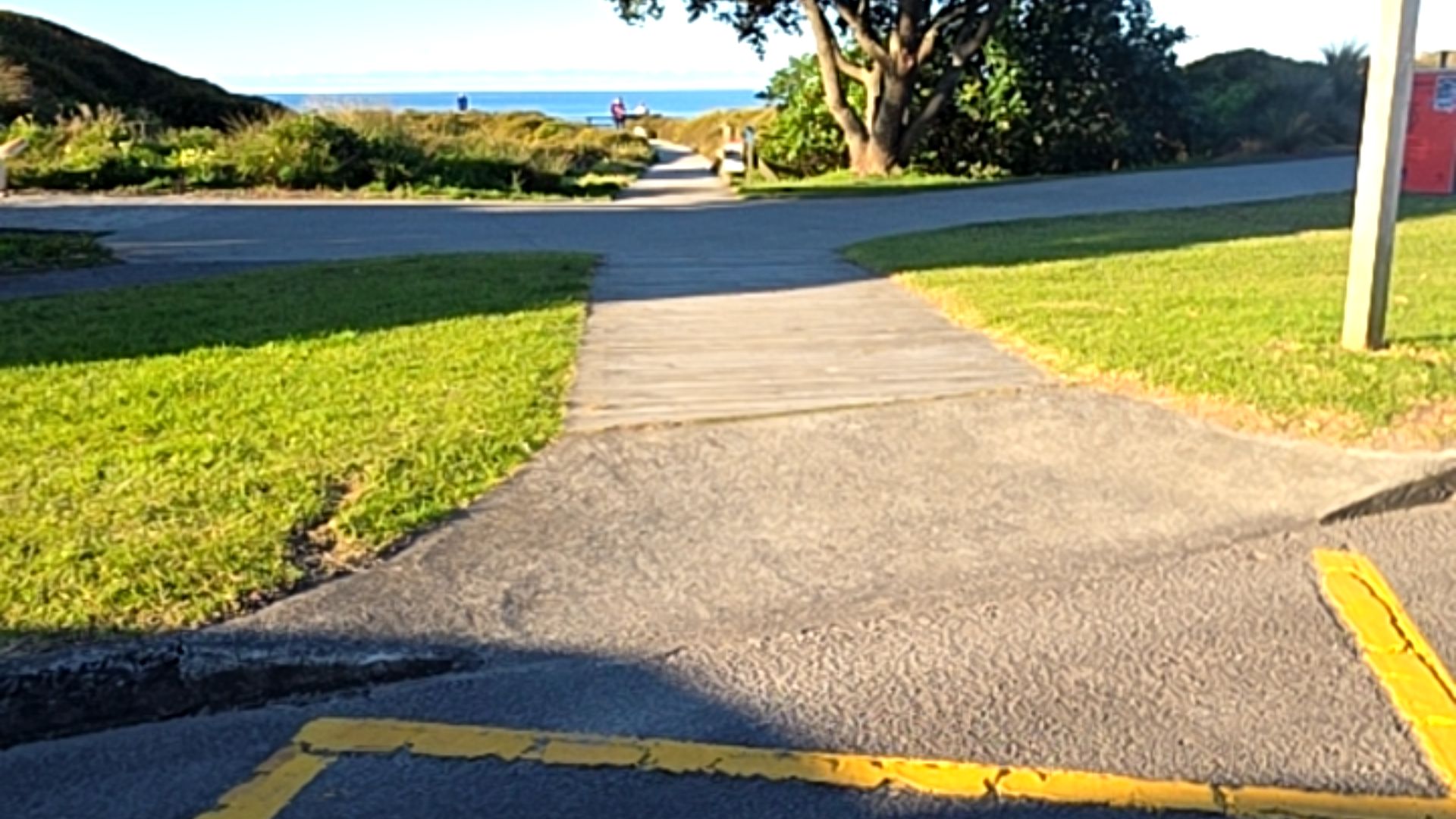 Wide asphalt path leads to a level concrete ramp and horizontal wooden plank boardwalk. The firm-surface pathway is bordered by grass and leads through coastal vegetation towards the ocean.