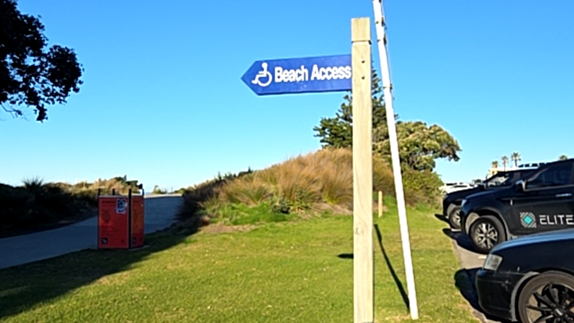 Blue arrow sign with white wheelchair icon and 'Beach Access' text points left to a level bitumen path. Timber post on grass verge next to a bitumen car park with parked black vehicles.