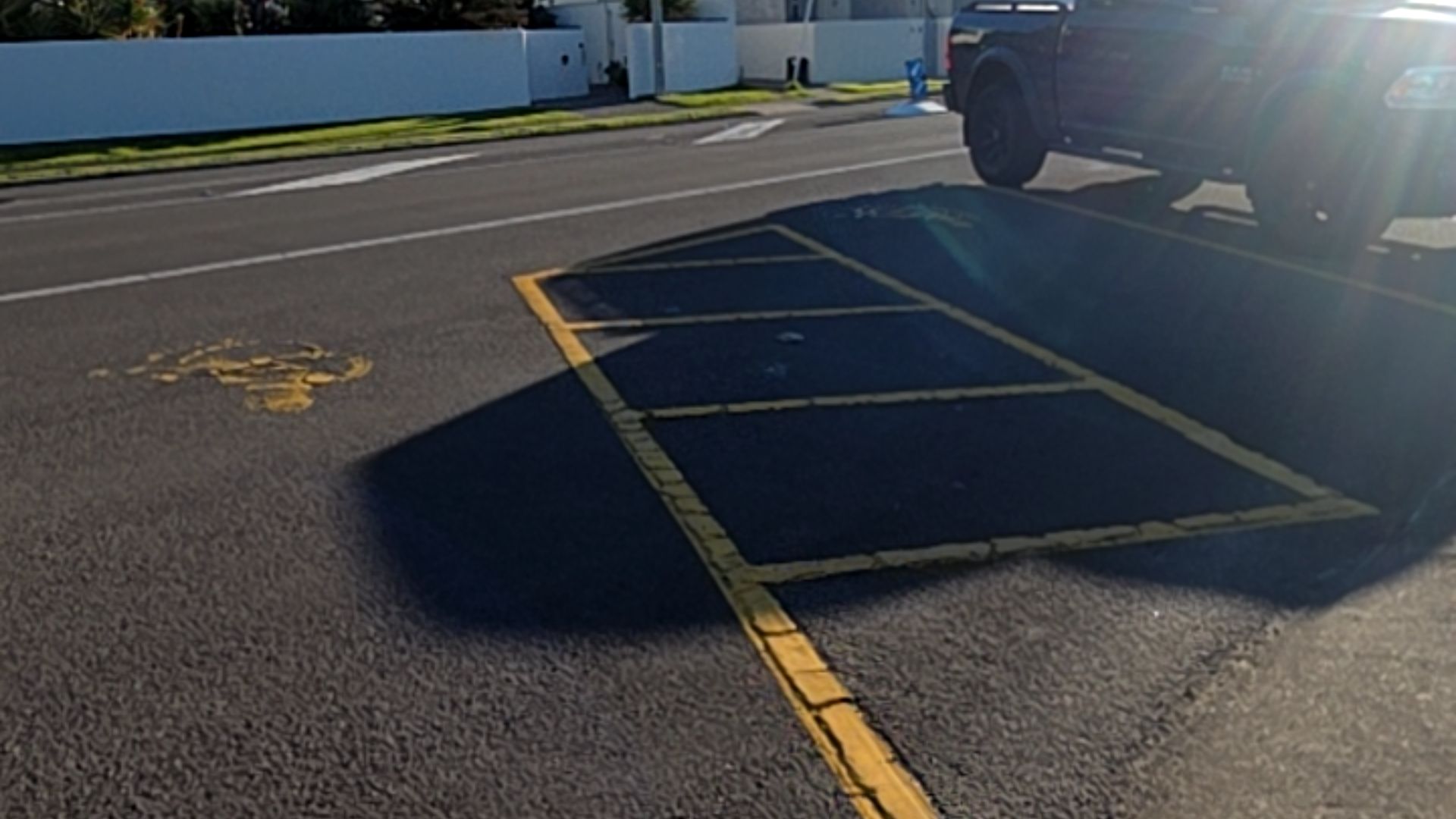 Yellow-lined parking bays on a level asphalt surface with a faded yellow accessibility symbol. A black ute is parked adjacent, casting a long shadow across the bitumen.