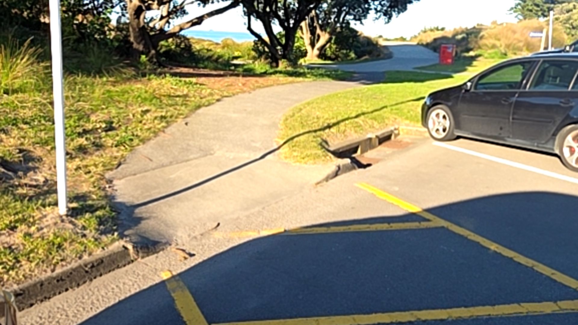 Concrete ramp with a slightly uneven transition from an asphalt car park with yellow markings to a paved pathway curving through parkland.