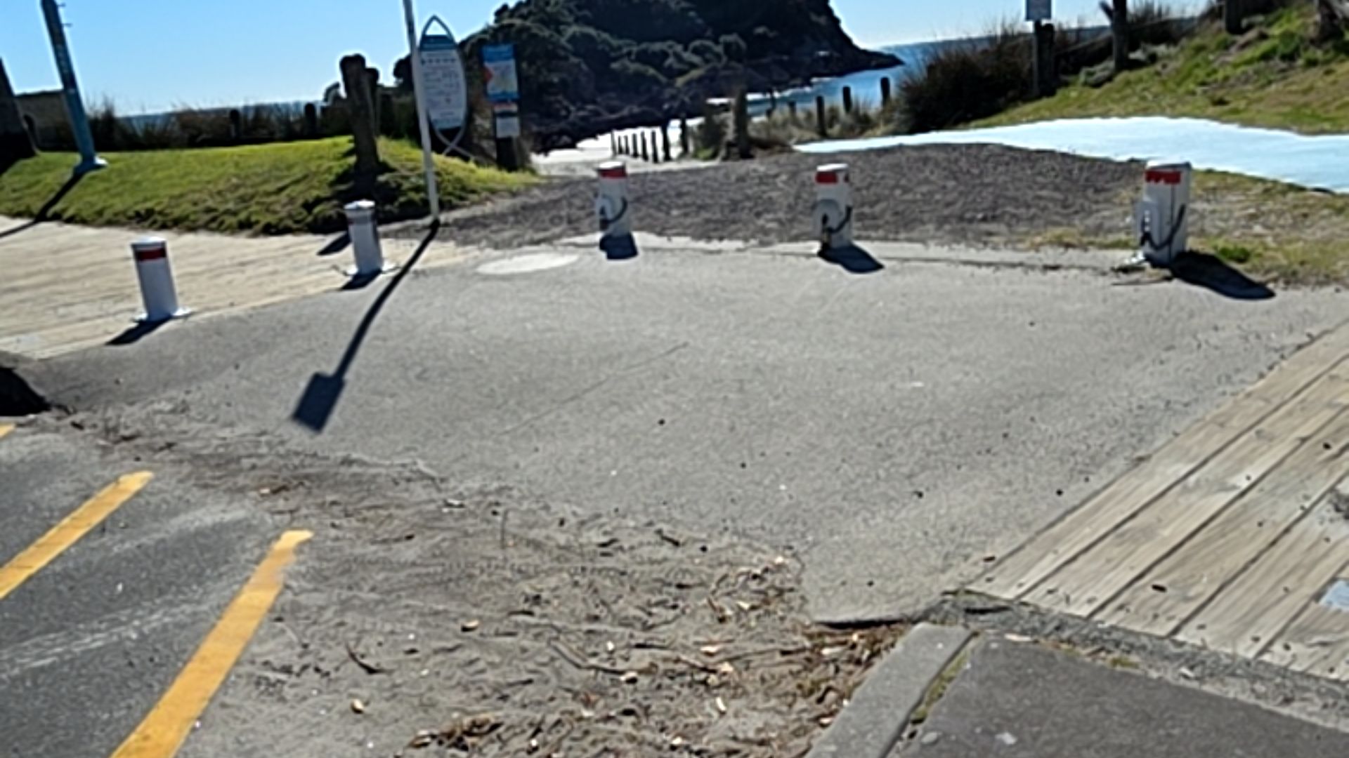 Level asphalt area with yellow diagonal markings beside a wooden plank walkway. Four white bollards with red tops are spaced across the flat path leading toward a grassy coastal area.