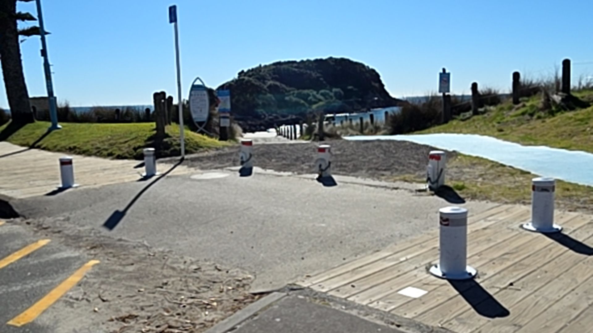 Level concrete area with yellow parking lines and white bollards transitions to a gravel path. A timber boardwalk and a blue paved path lead towards the coast and a large rocky hill.
