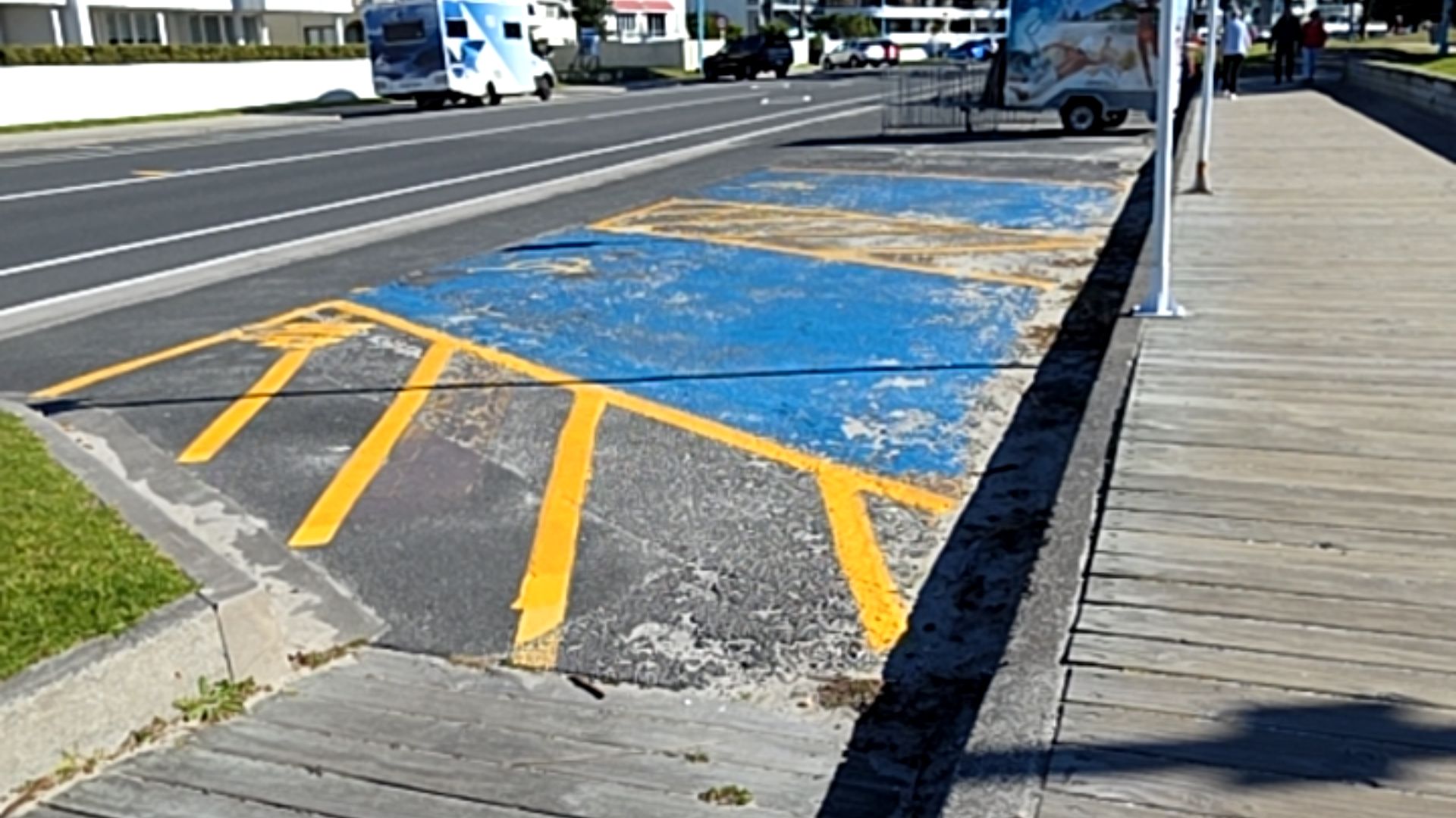 Level accessible parking space with faded blue paint and yellow hatched markings on bitumen. It sits directly adjacent to a grey wooden boardwalk and a two-lane bitumen road.