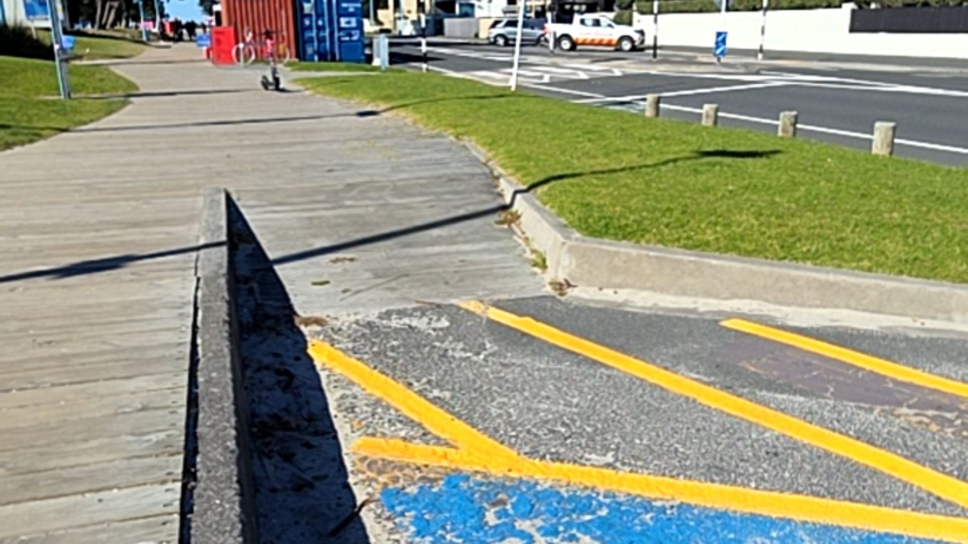 Wide level timber boardwalk transitions via a concrete kerb ramp to asphalt with high-visibility yellow markings and blue paint. A low concrete wall borders the path to the left.