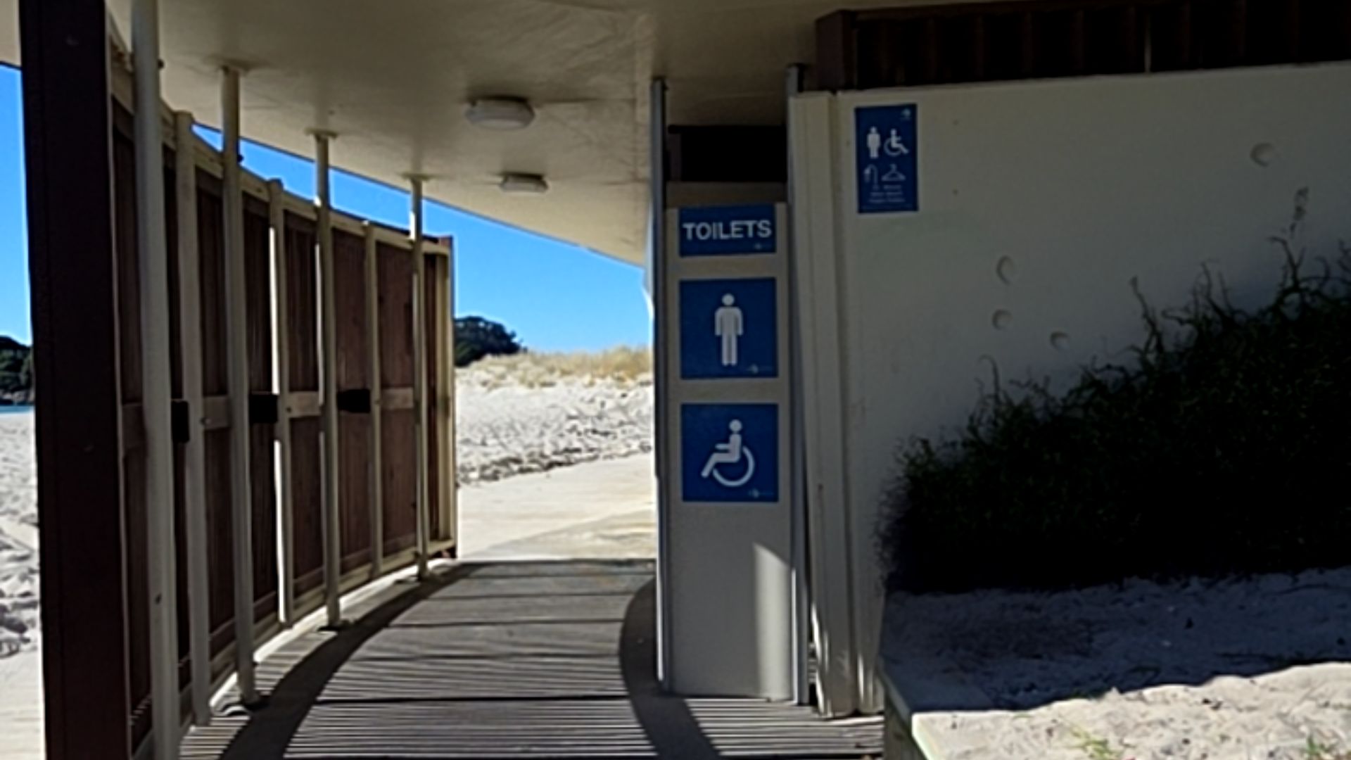 Level wooden slatted boardwalk leads past a white pillar with blue signs for male and wheelchair accessible toilets. Undercover area with flat floor surfaces and clear signage.
