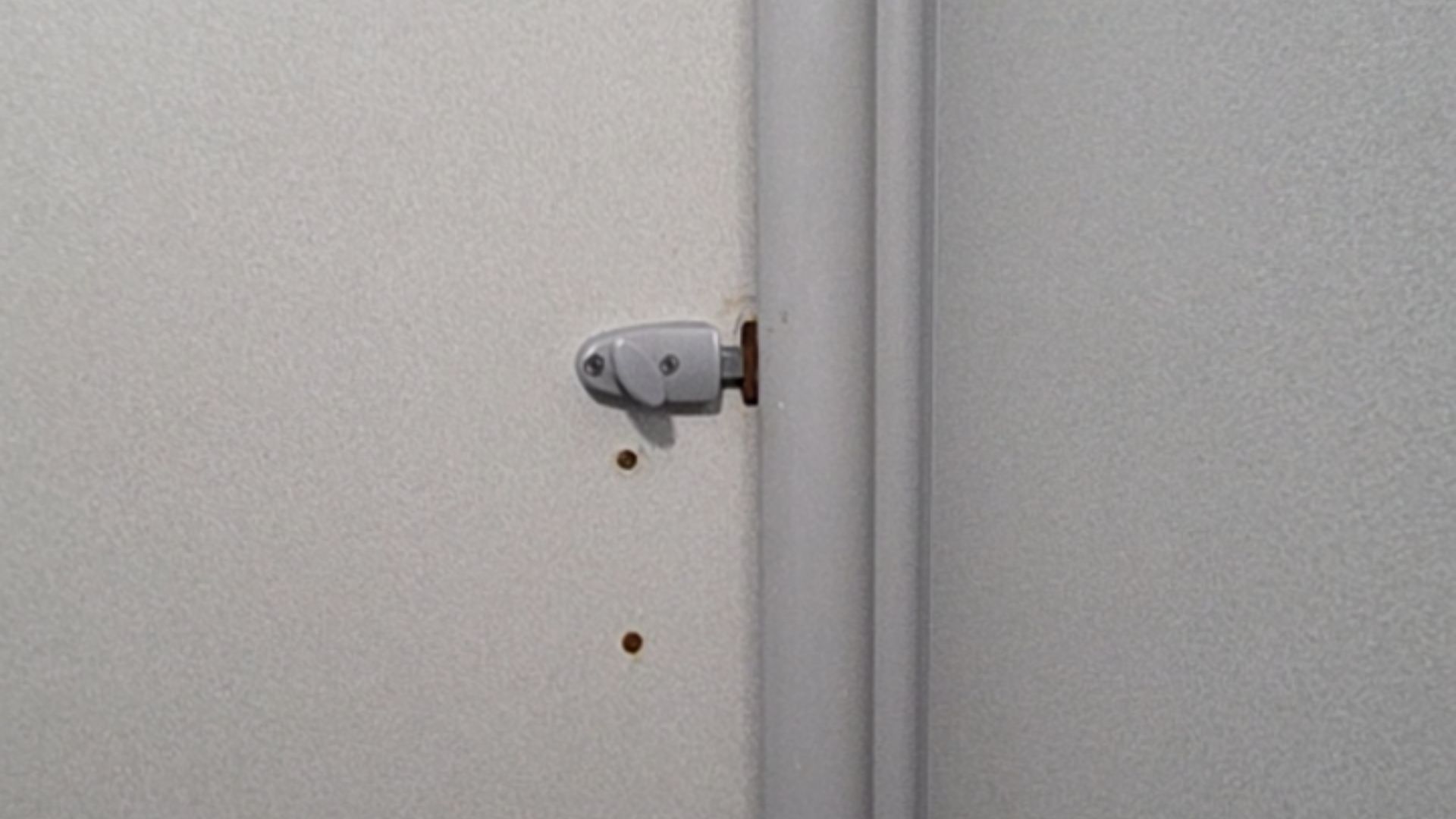 Small grey thumb-turn lock on a light grey textured door, adjacent to a vertical door frame. Two small screw holes are visible on the door surface below the lock mechanism.