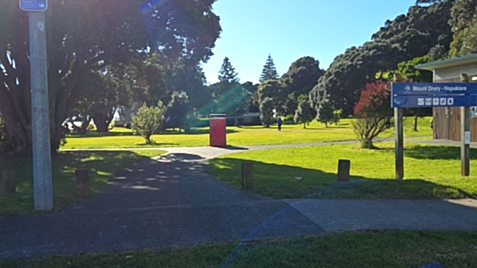 Level asphalt pathway through a grassy park. Blue sign on wooden posts identifies "Mount Drury - Hopukiore" with white icons. Small wooden bollards and a red bin are located along the path.