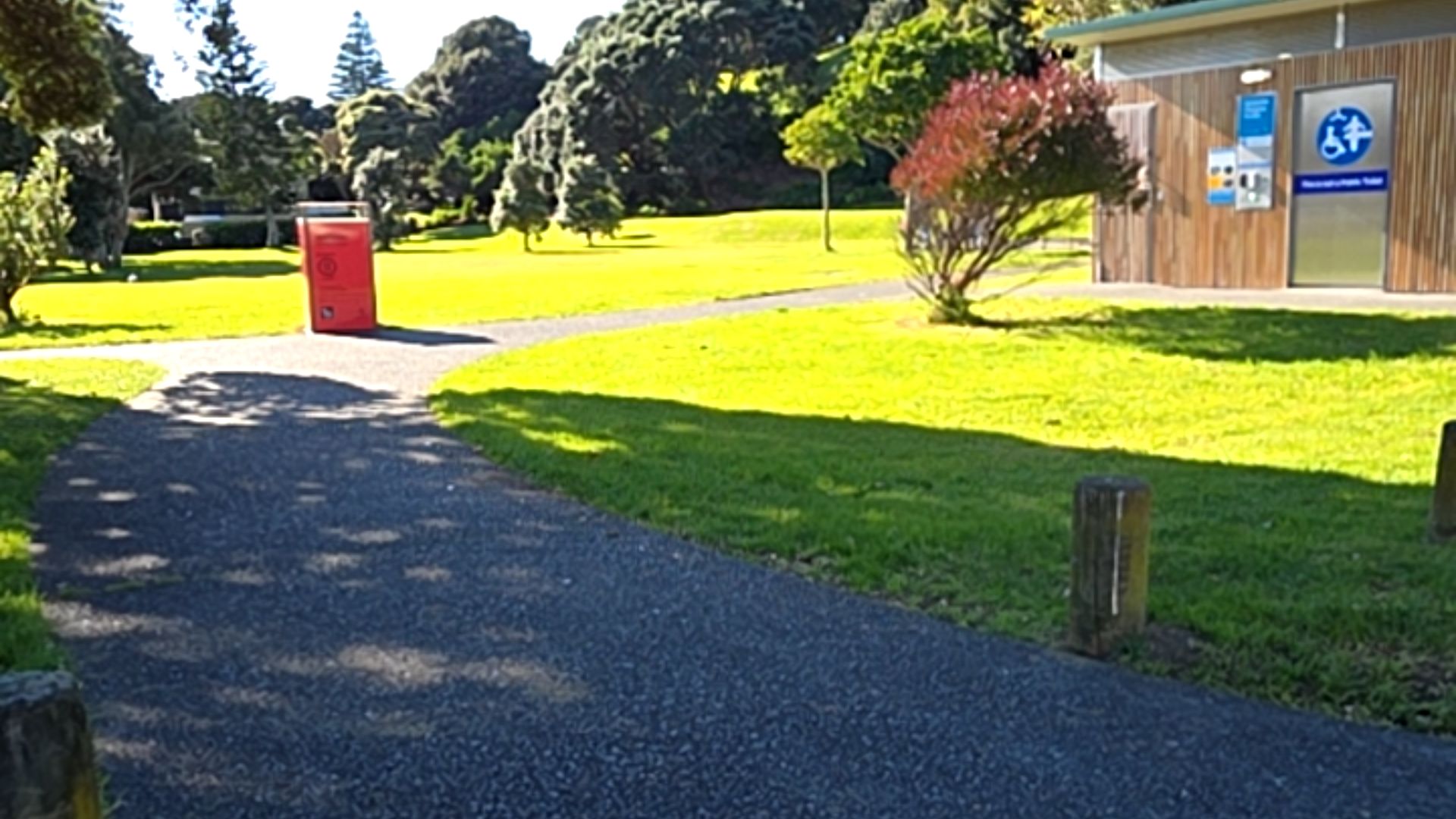 Wide level asphalt pathway leads to a wooden building with a silver door featuring a blue accessibility symbol. Set in a grassy parkland with trees and a red bin.