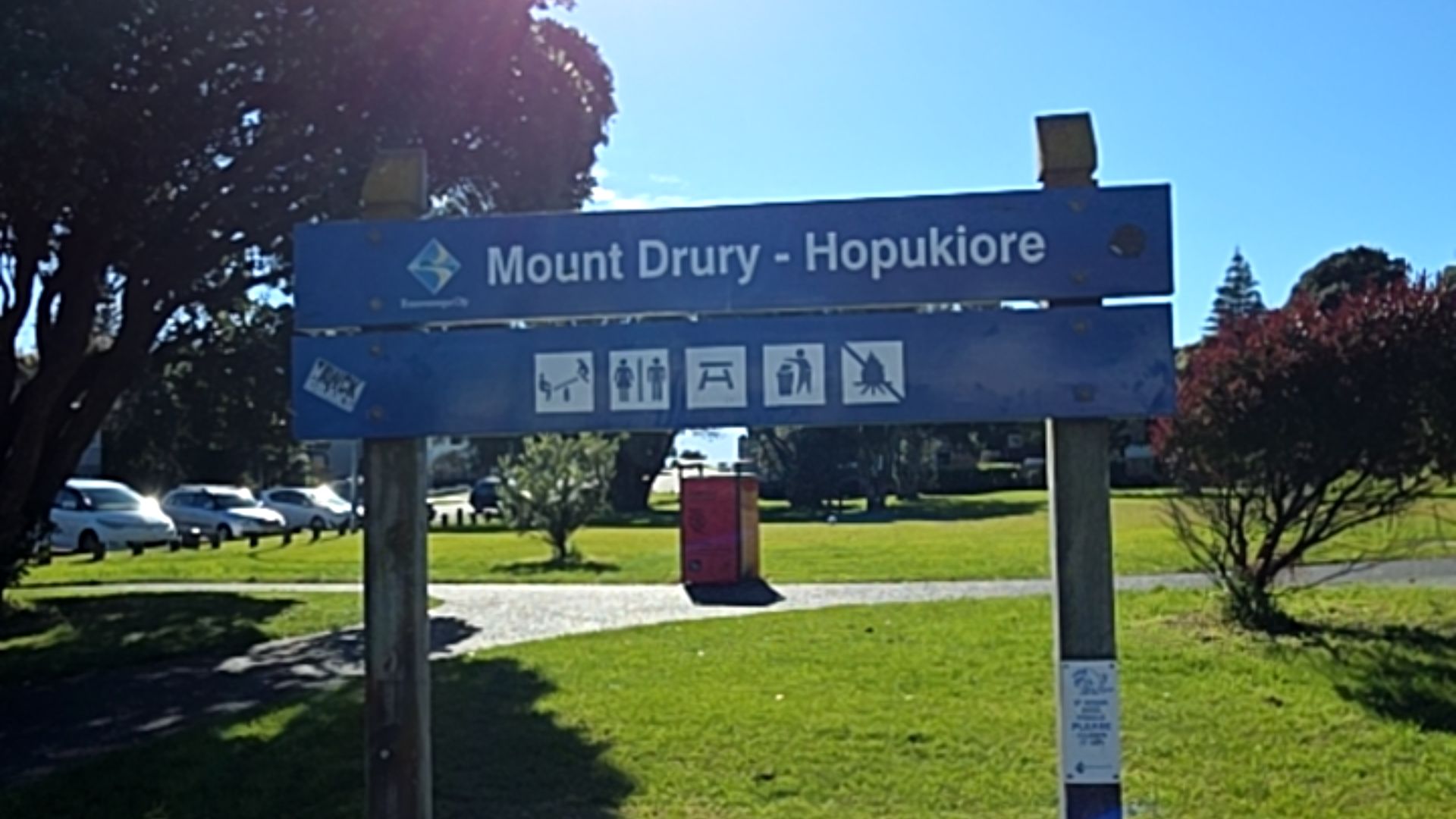 Blue park sign "Mount Drury - Hopukiore" with icons for toilets, playground, and picnic area. It is set on wooden posts beside a level concrete pathway through flat grassed parkland.