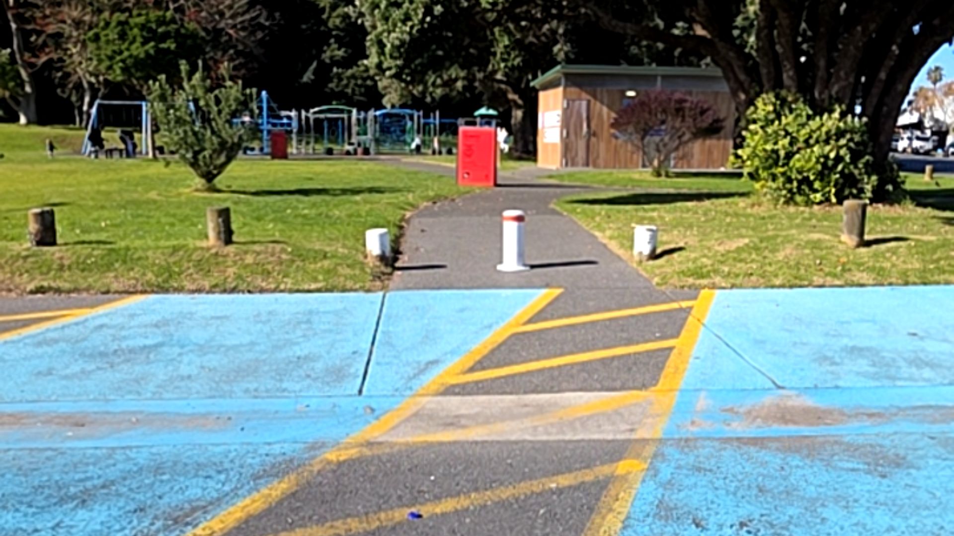 Level asphalt path from blue parking with yellow hatching. Three white bollards at entrance, one in the centre with red stripe. Grassy park, playground, and timber building in background.