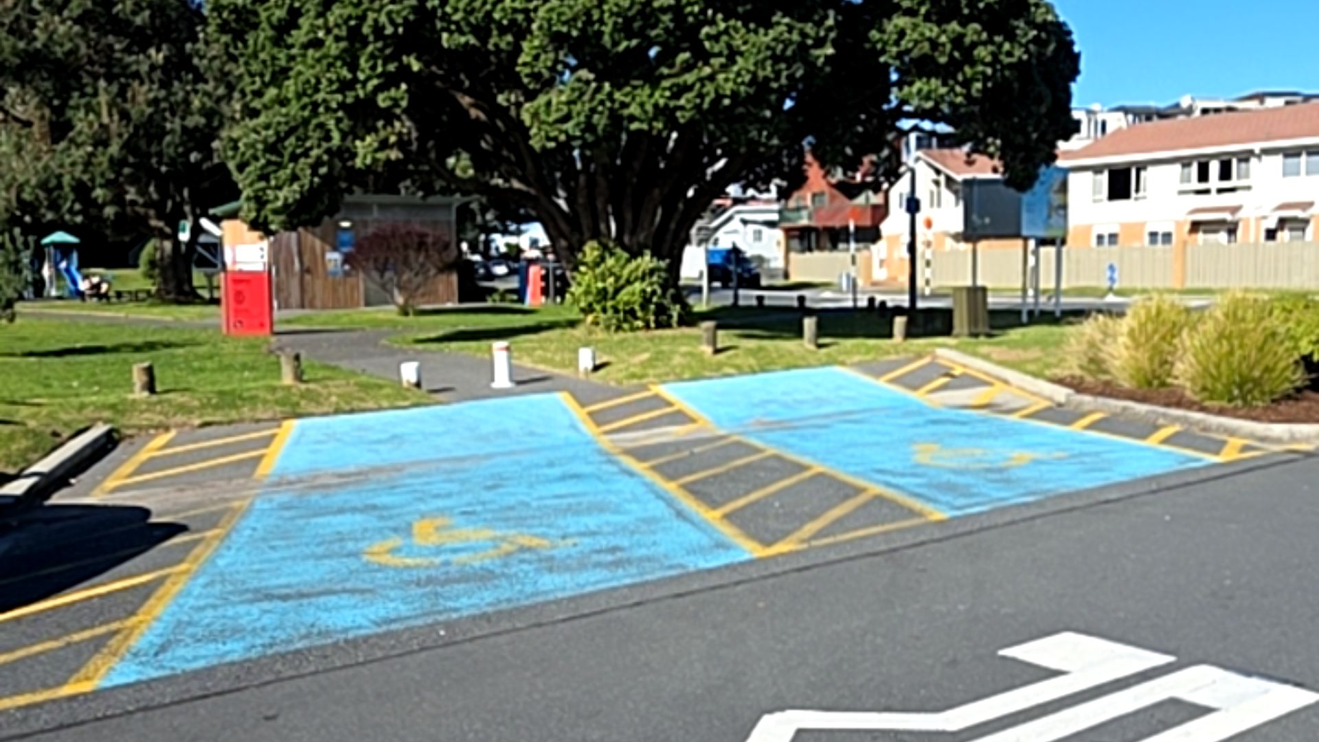 Two accessible parking spaces with blue surfaces and yellow wheelchair symbols. Yellow hatched access zones are provided. Level asphalt surface leads to a pathway and grassy park area.