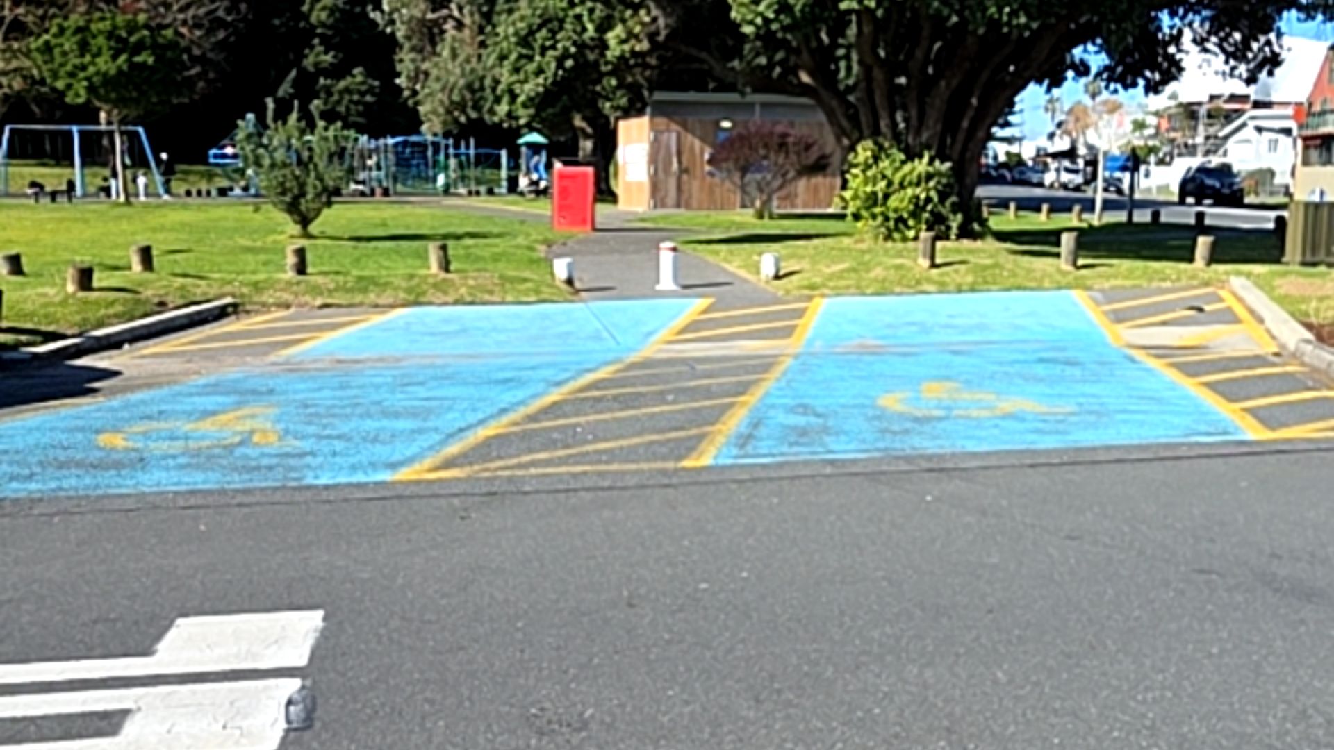 Two blue accessible parking spaces with yellow wheelchair symbols and hatched markings on level bitumen. A level path with bollards leads from the spaces into a park with a playground.