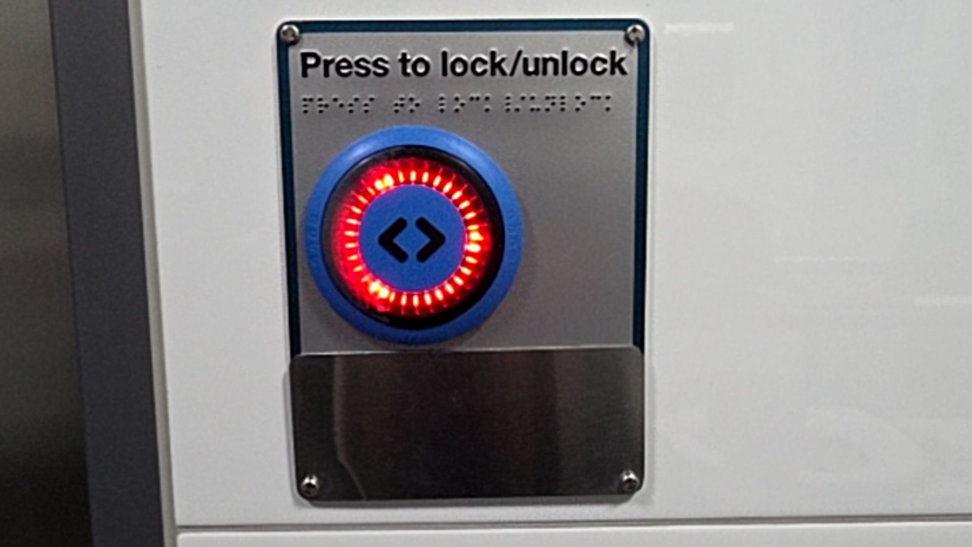 Silver-coloured door control panel with 'Press to lock/unlock' text and tactile Braille. A blue button in the centre has a red illuminated LED ring and black door symbols.