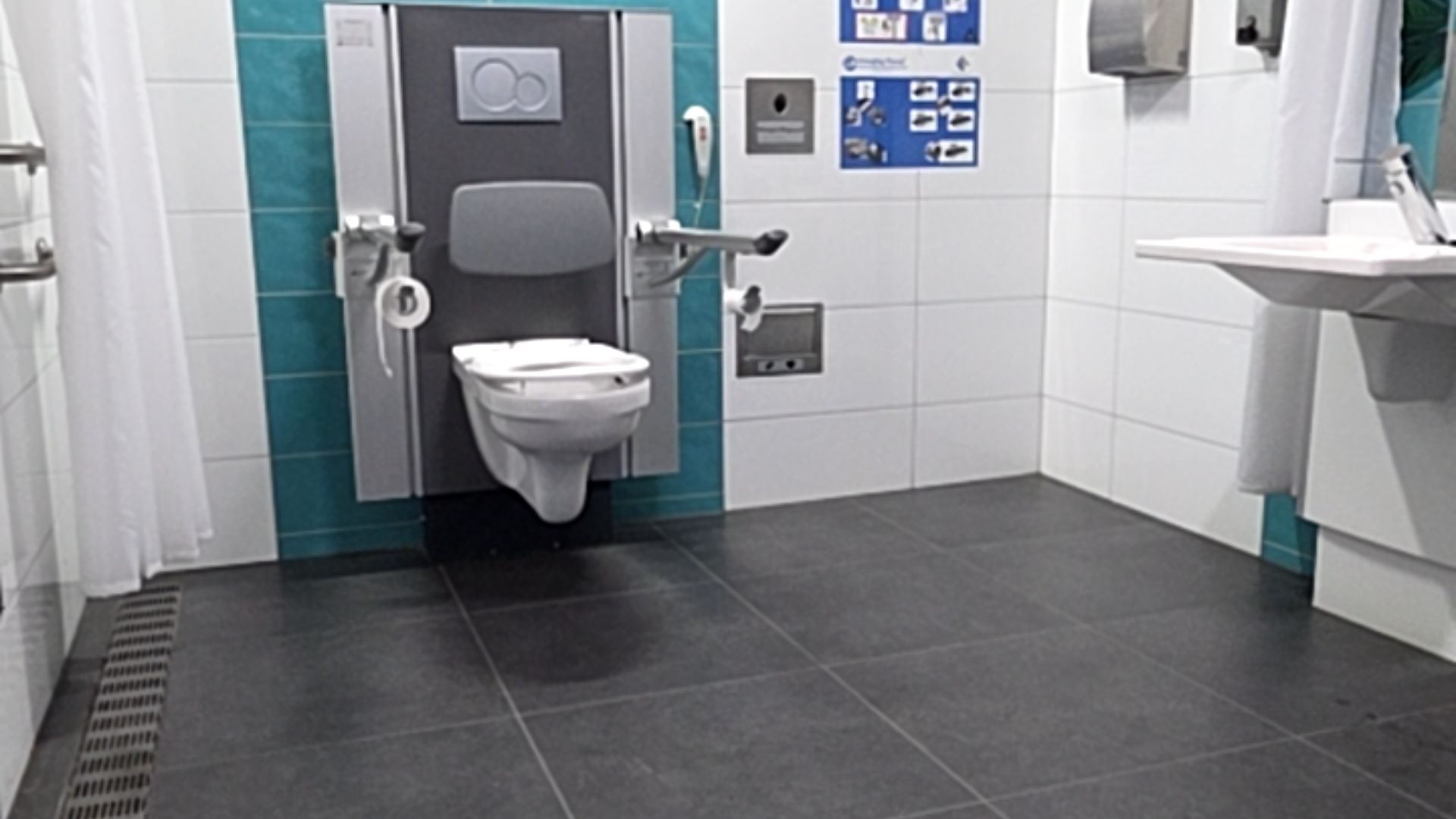 Wall-hung toilet with grey backrest and swing-away grab bars. Level dark grey tiled floor. Wall-mounted sink, emergency cord, and shower area with floor-level drain and white curtain.