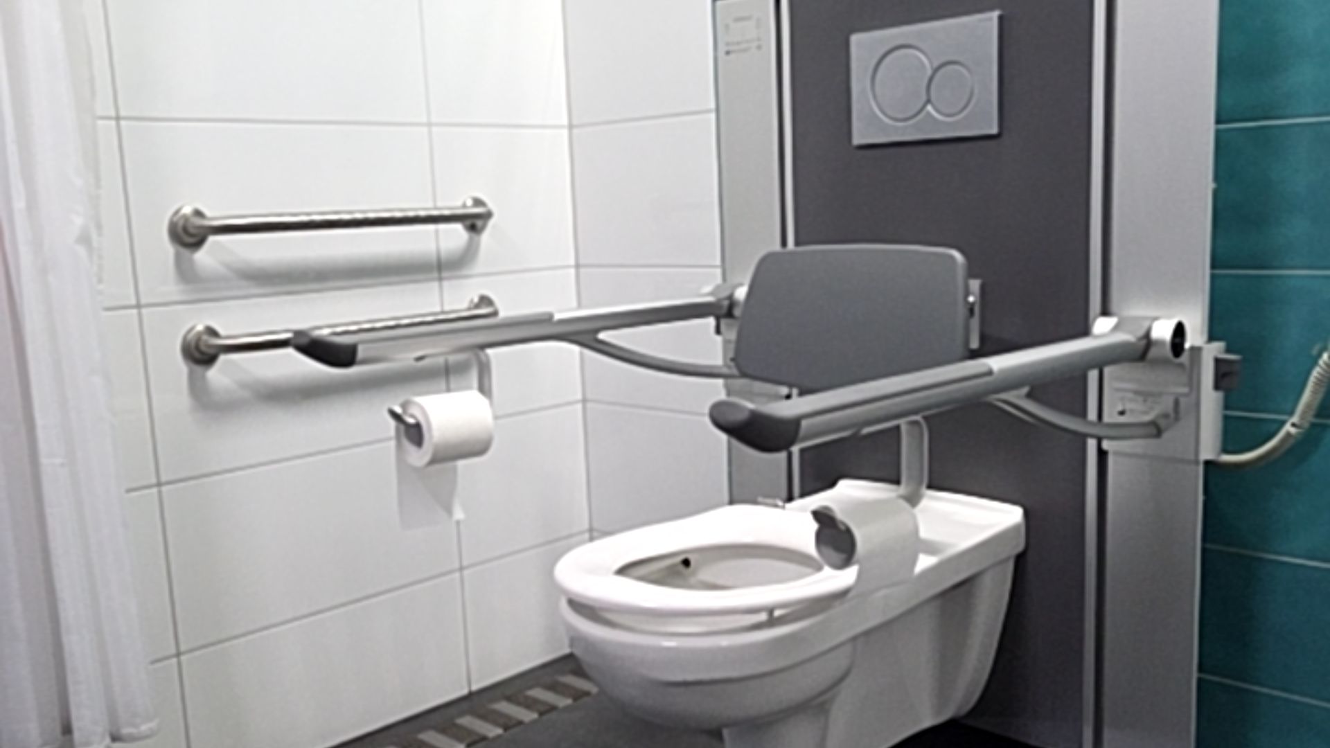 Wall-mounted toilet with padded backrest and two flip-down armrests. Two horizontal stainless steel grab bars on white tiled wall. Large grey dual flush button and non-slip floor tiles.